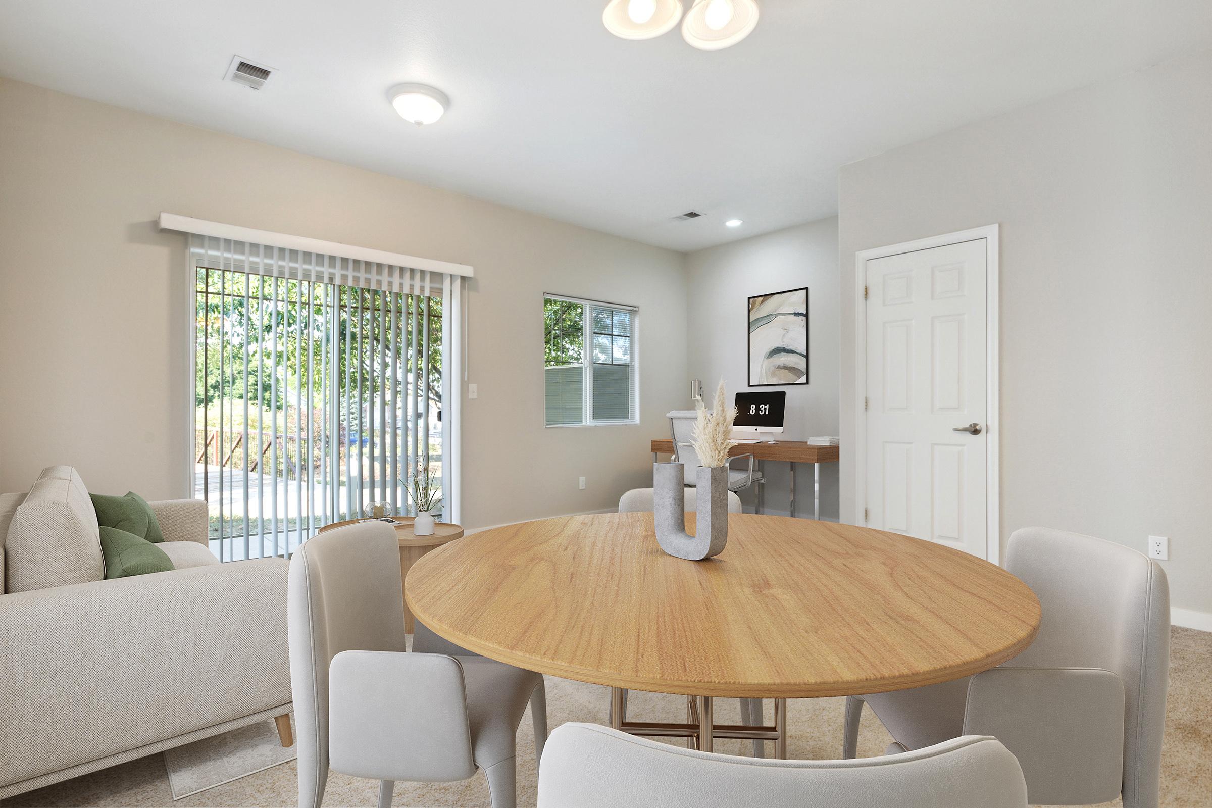 A cozy living area featuring a round wooden dining table with light gray chairs. In the background, a beige couch with green cushions and a window with blinds. A small desk with a computer can be seen, along with light-colored walls and soft carpet, creating a warm and inviting atmosphere.
