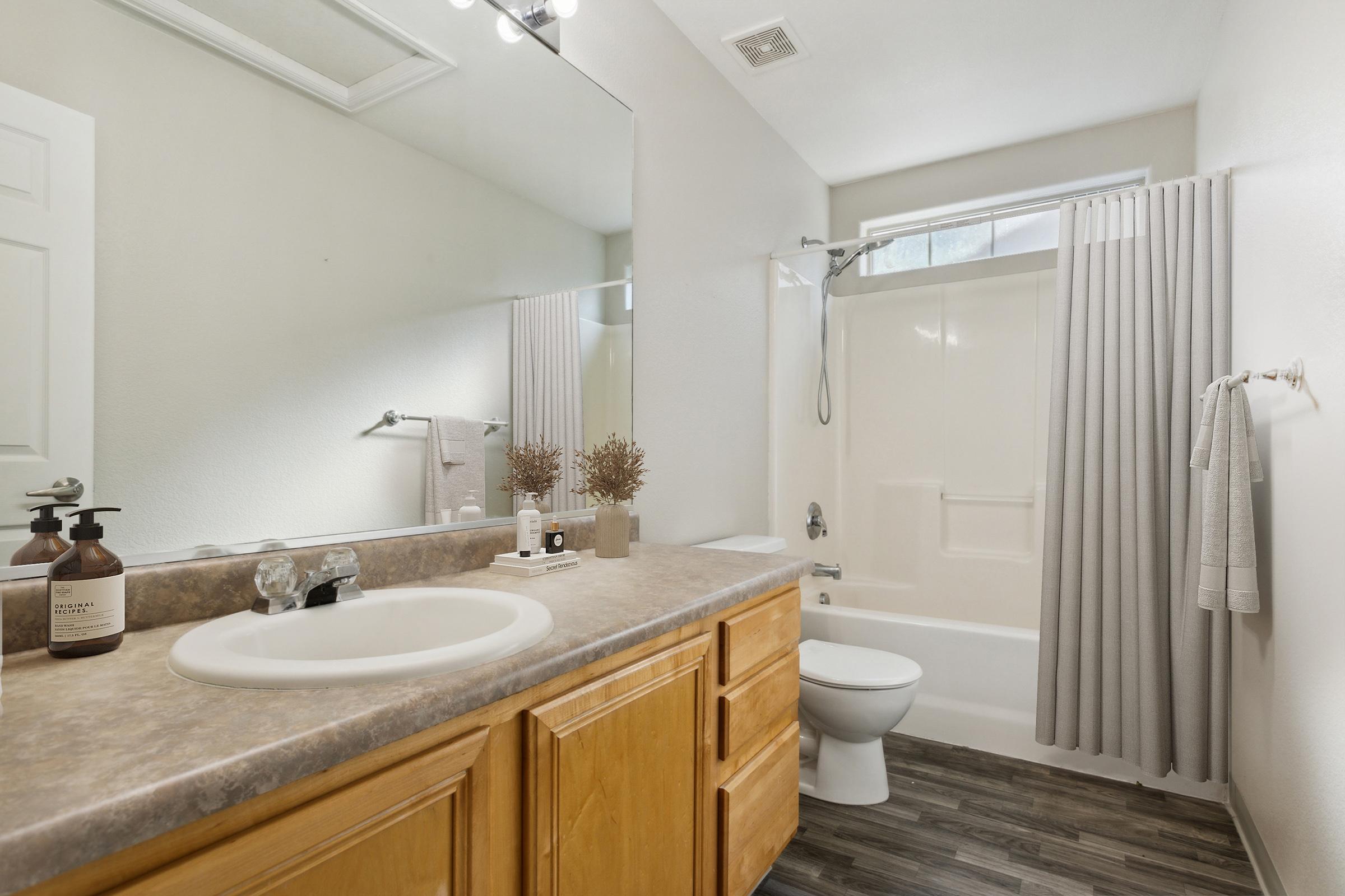 A clean and modern bathroom featuring a bathtub with a shower, a wooden vanity with a sink, and a large mirror. The space has light-colored walls and wooden flooring, with neutral curtains and simple decor items on the vanity, creating a fresh and inviting atmosphere.