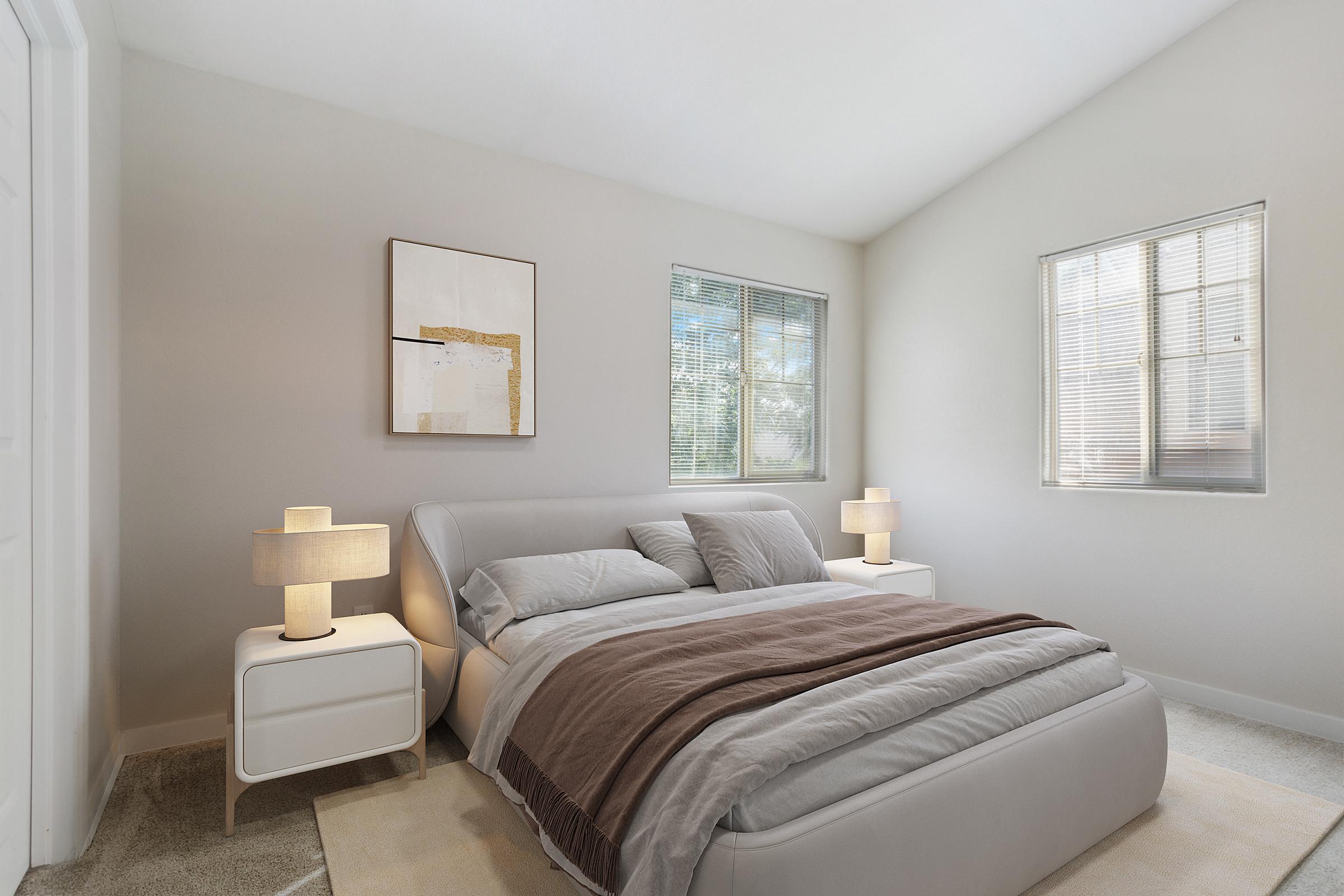 A modern bedroom featuring a plush bed with soft bedding, two bedside tables with stylish lamps, and large windows allowing natural light. The walls are painted in a light color, and there is a minimalist piece of artwork above the bed, creating a serene and inviting atmosphere.
