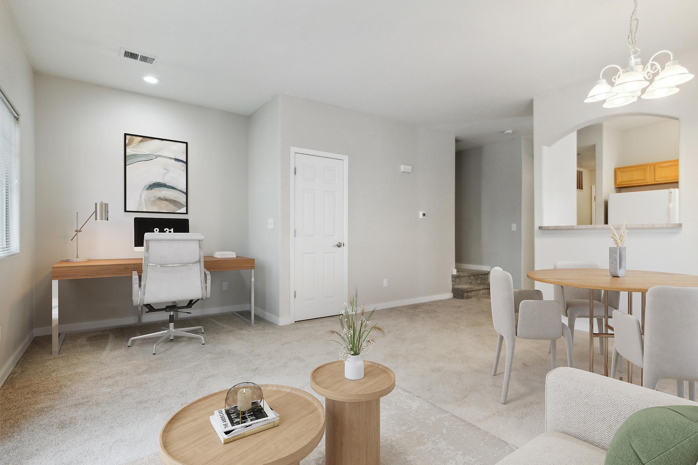 A modern living room featuring a light-colored carpet, a small wooden desk with a chair, and a round dining table with gray chairs. There is a decorative plant on the table, a stylish coffee table with a glass centerpiece, and a wall art piece above the desk. The space is bright and airy with soft lighting.