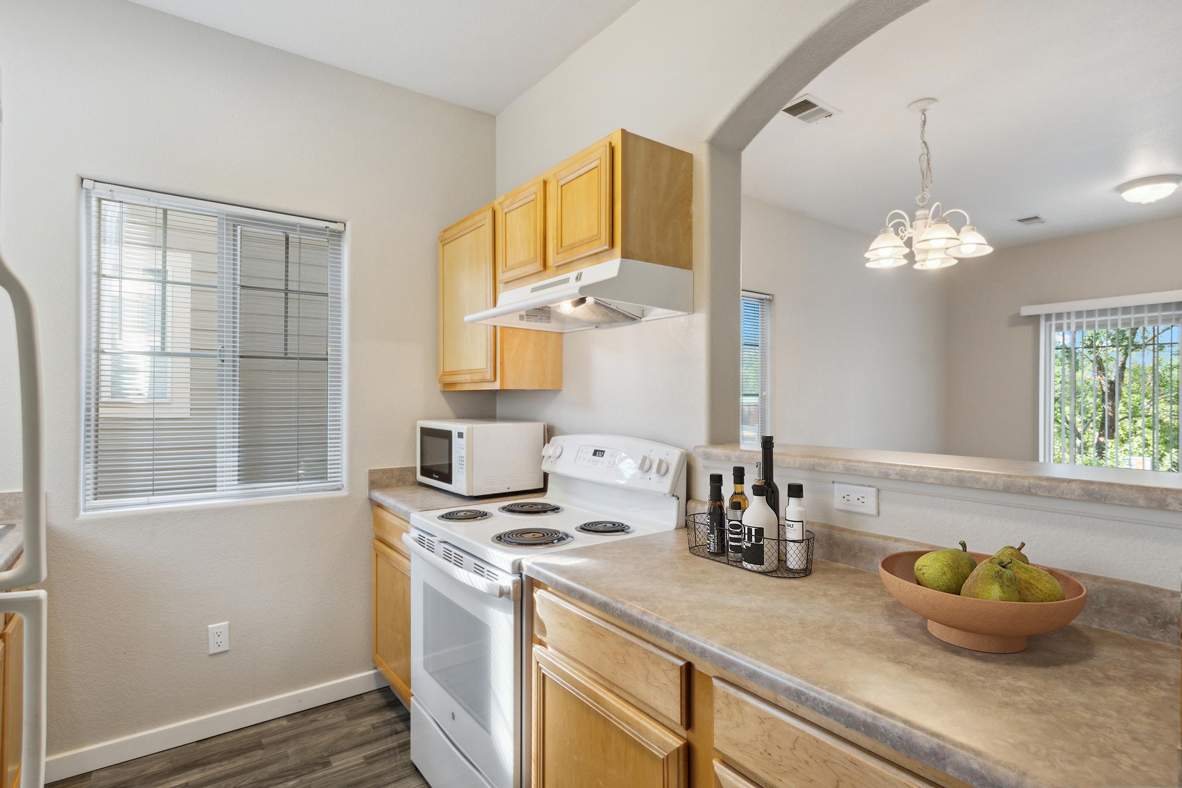 A modern kitchen featuring a white stove, microwave, and wooden cabinets. The countertop holds a bowl of green pears. Natural light comes through a nearby window with blinds, illuminating the space. A light fixture hangs above, adding warmth to the room's neutral tones.