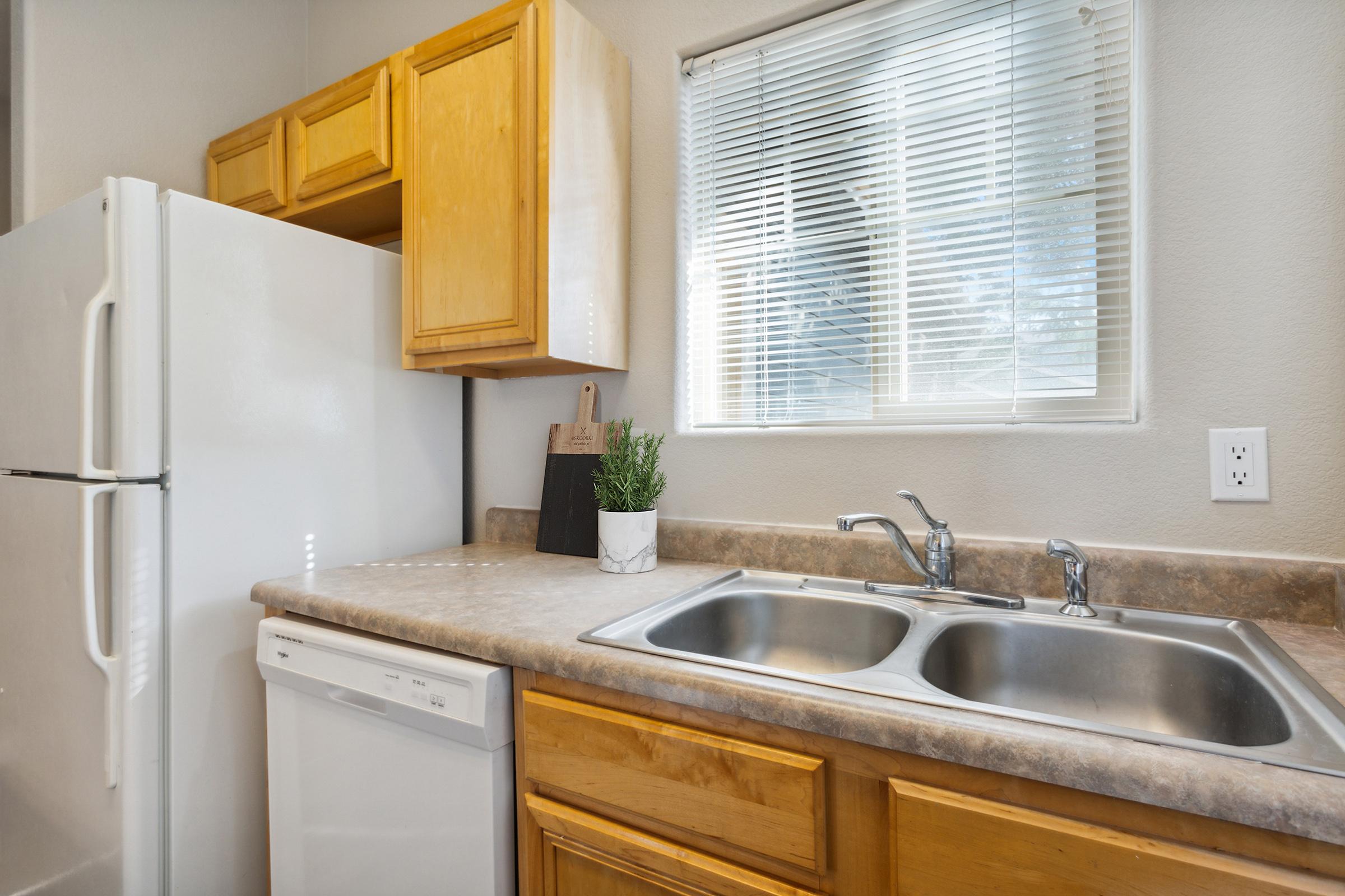 A modern kitchen features light-colored wooden cabinets, a double sink, and a white dishwasher. A refrigerator stands to the left, and a window allows natural light to enter. A small plant is placed on the countertop, enhancing the inviting atmosphere.