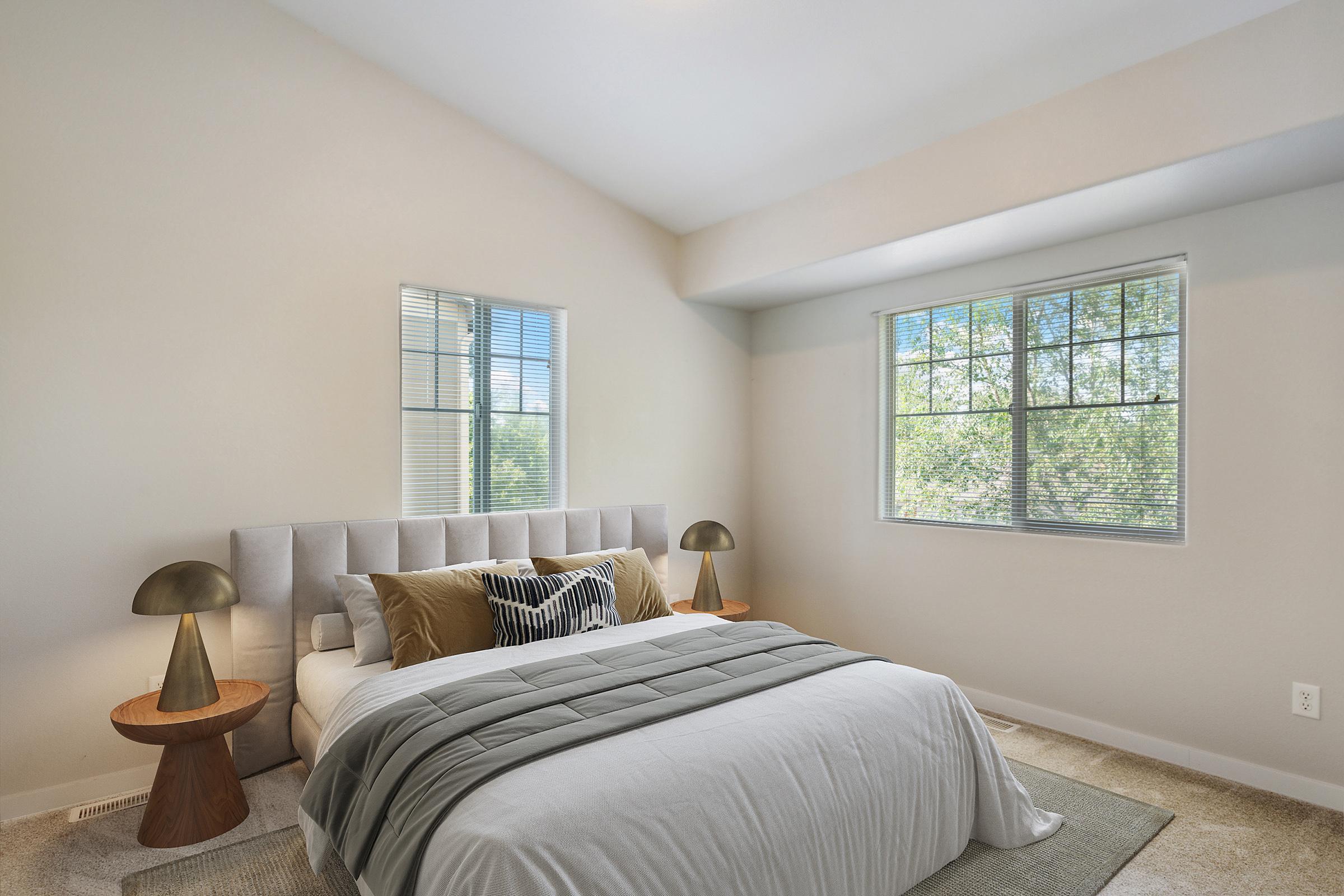 A cozy, well-lit bedroom featuring a light-colored bed with decorative pillows, two modern bedside tables with lamps, and large windows that allow natural light. The walls are painted in a soft, neutral tone, and a plush rug is placed on the carpeted floor, creating a tranquil atmosphere.