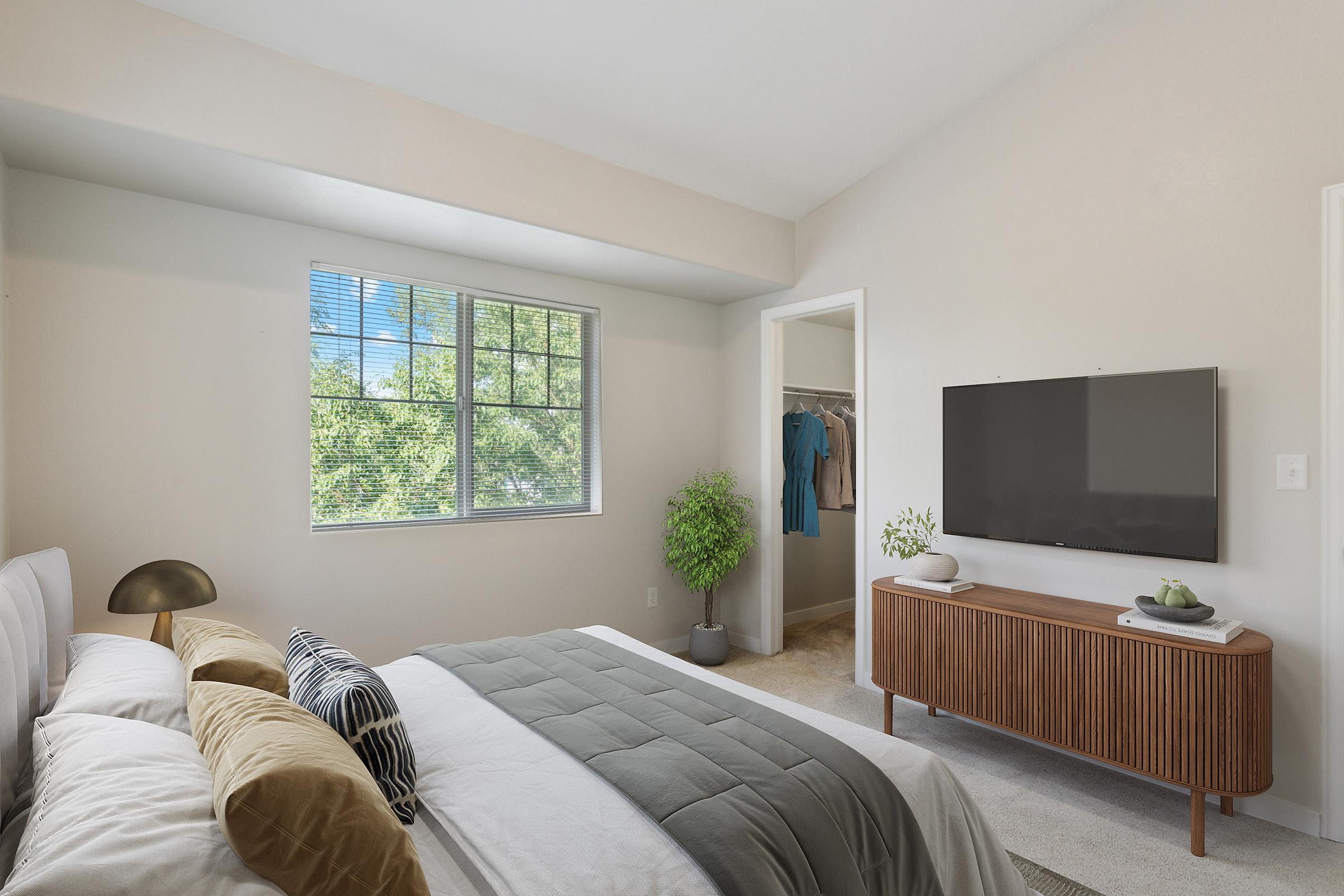 A cozy bedroom featuring a comfortable bed with decorative pillows, a wooden dresser with a TV mounted above, and a window letting in natural light. There's a small plant on the dresser and a closet visible in the background. The walls are painted light colors, creating a serene atmosphere.