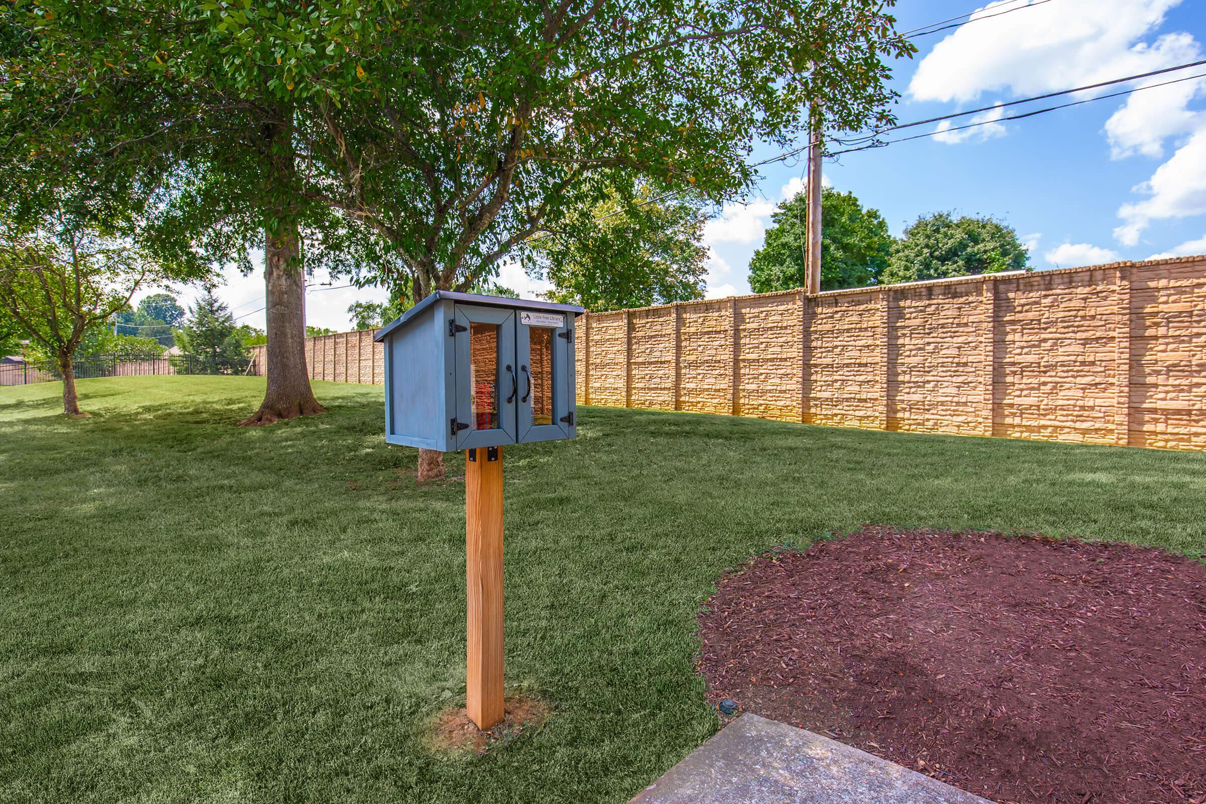 A small blue wooden library box on a post in a grassy yard, surrounded by trees and a stone wall. The sky is bright with fluffy clouds. The area is well-maintained, featuring a circular mulch bed nearby.