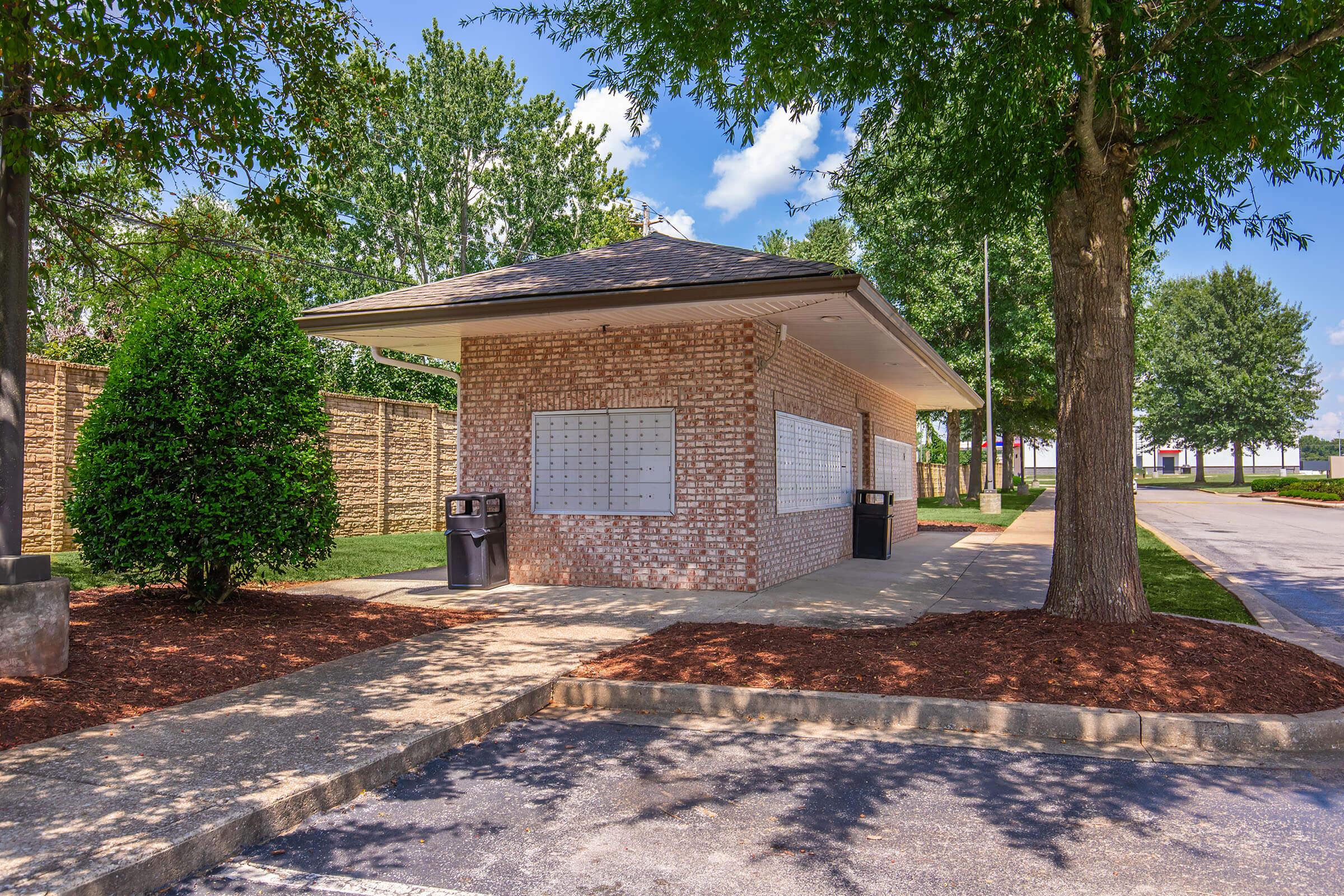 A small brick building with a dark roof and multiple mailboxes on the exterior. The structure is surrounded by trees and shrubs, with two trash bins nearby. The pathway leading to the building is clear, and there are no vehicles in sight. The area is well-lit with blue skies and fluffy clouds above.