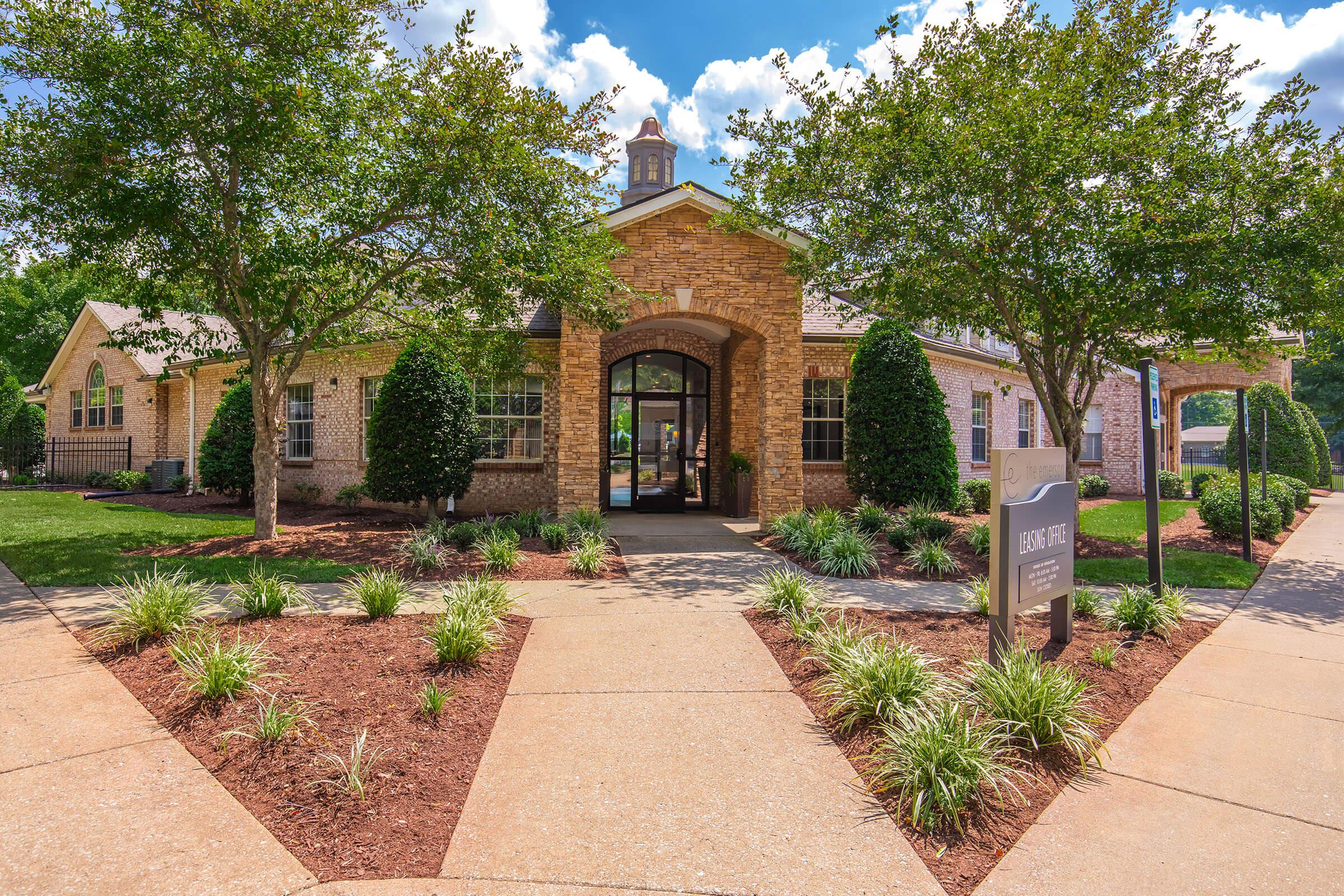 A welcoming brick building with a large entrance, surrounded by well-maintained landscaping featuring green grass, small bushes, and trees. A pathway leads to the entrance, and a sign is visible near the front. The sky is clear with a few fluffy clouds.