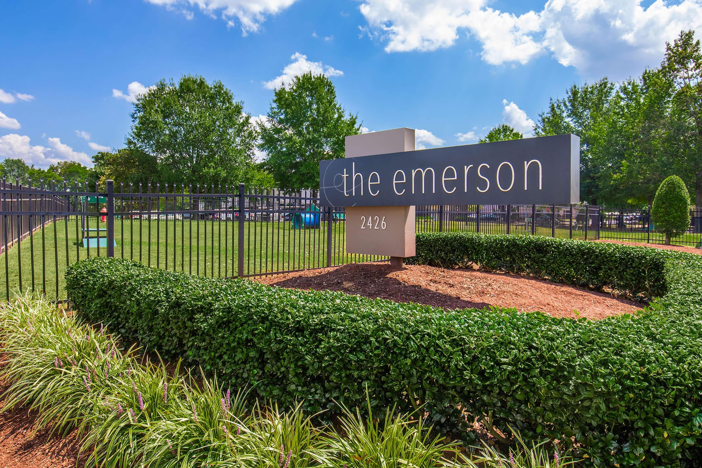 A modern sign for "the emerson" located at 2426, set against a backdrop of a well-maintained green area with trees and shrubs. The sky is partly cloudy, adding to the bright and inviting atmosphere of the location. The fence surrounding the area is neatly arranged, enhancing the overall appeal of the site.