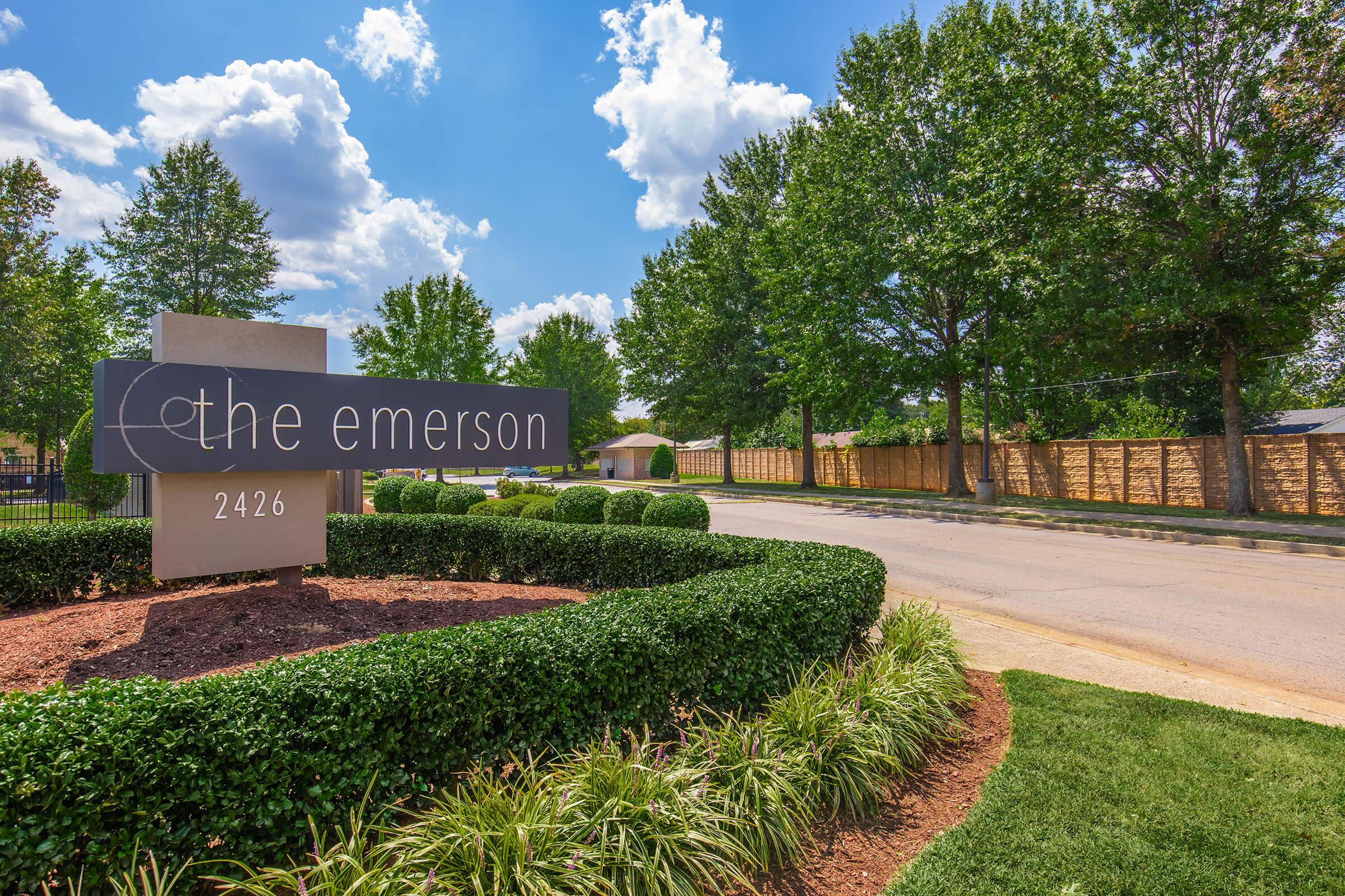 Sign for "The Emerson" located at 2426, surrounded by lush greenery and landscaping. The background features a clear blue sky with fluffy white clouds, and a well-maintained road lined by trees, creating a welcoming atmosphere.