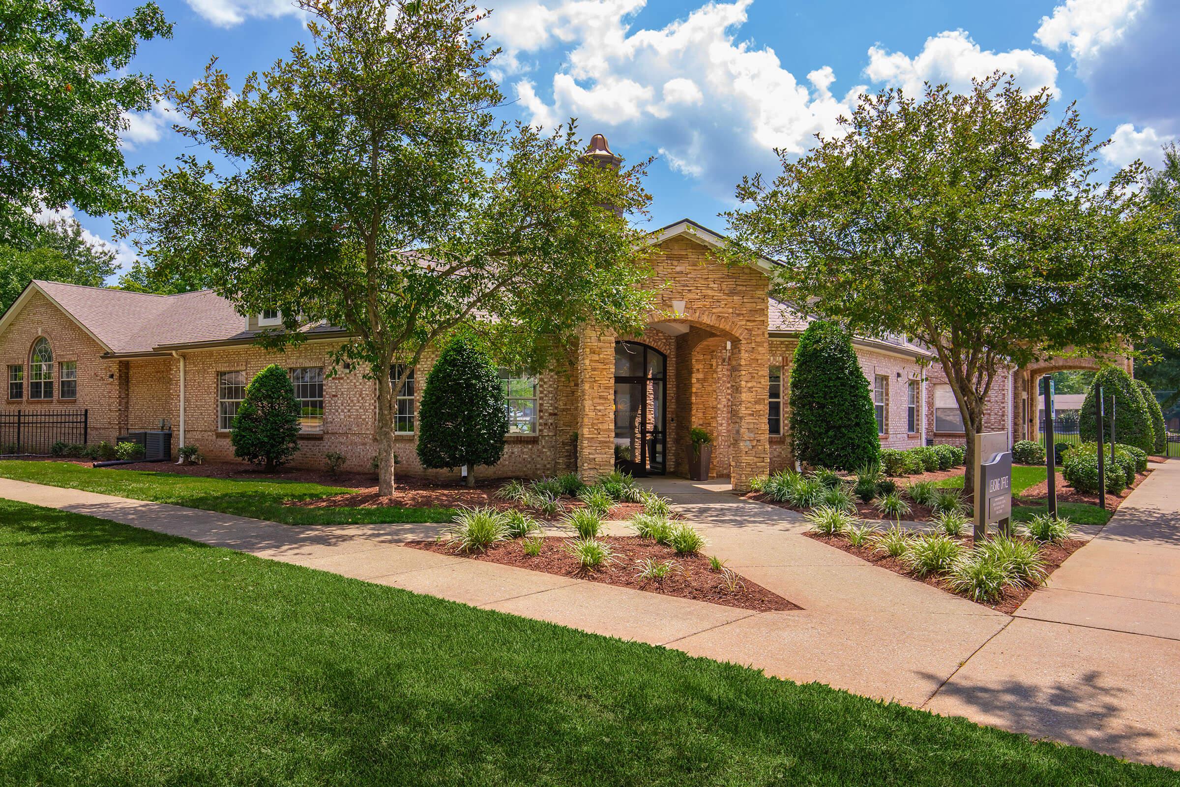 A well-maintained brick building surrounded by lush green lawns, shrubs, and trees under a blue sky with fluffy white clouds. The entrance features an archway and landscaped pathways. The setting conveys a welcoming and serene atmosphere, ideal for residential or community use.