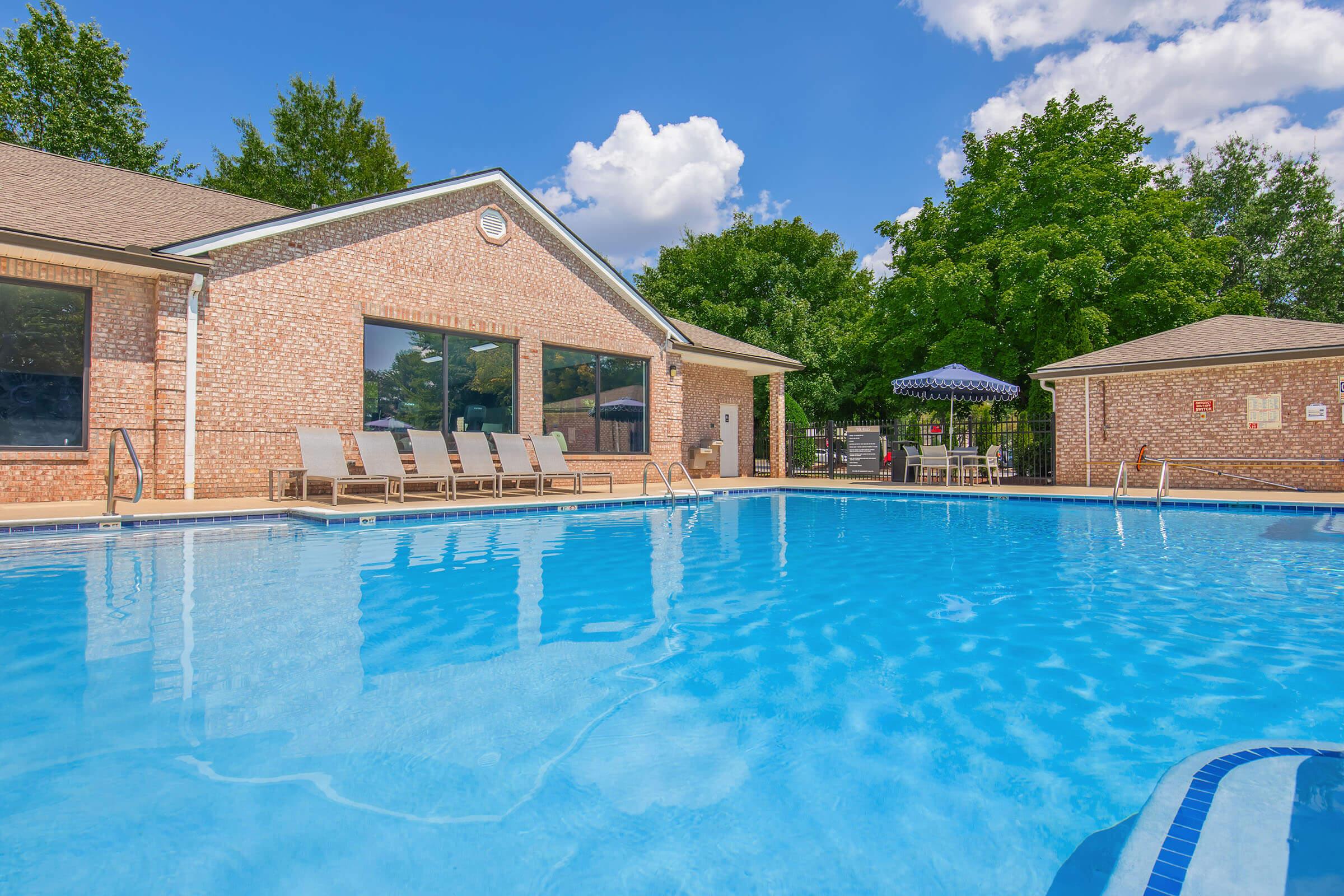 A clear blue swimming pool surrounded by lounge chairs and trees. In the background, there is a brick building with large windows and a shaded seating area under an umbrella. The sky is bright with a few fluffy clouds.