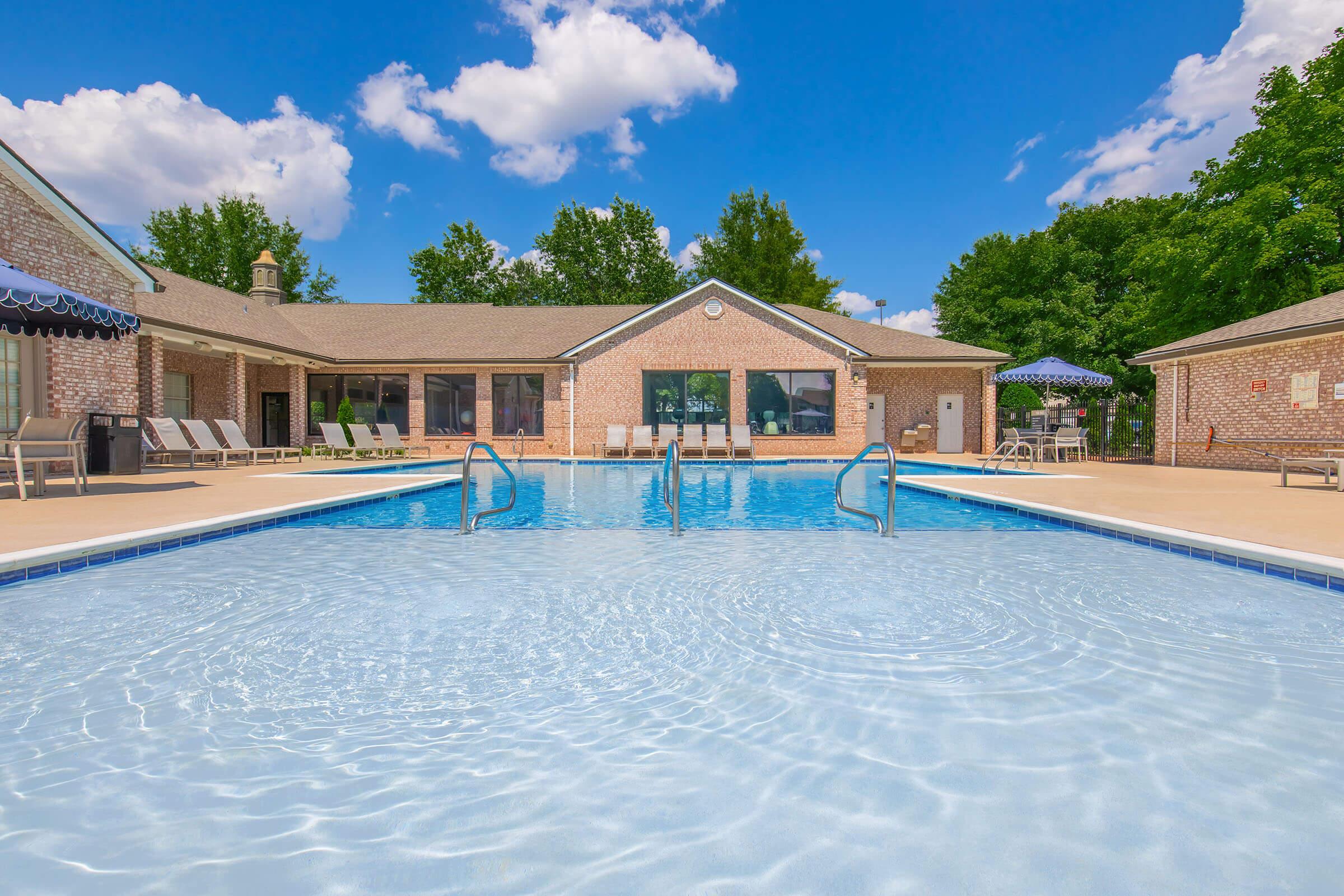 A clear blue swimming pool with steps leading in, surrounded by lounge chairs and a patio area. In the background, there is a single-story building with large windows and a treed landscape under a bright blue sky with fluffy white clouds.