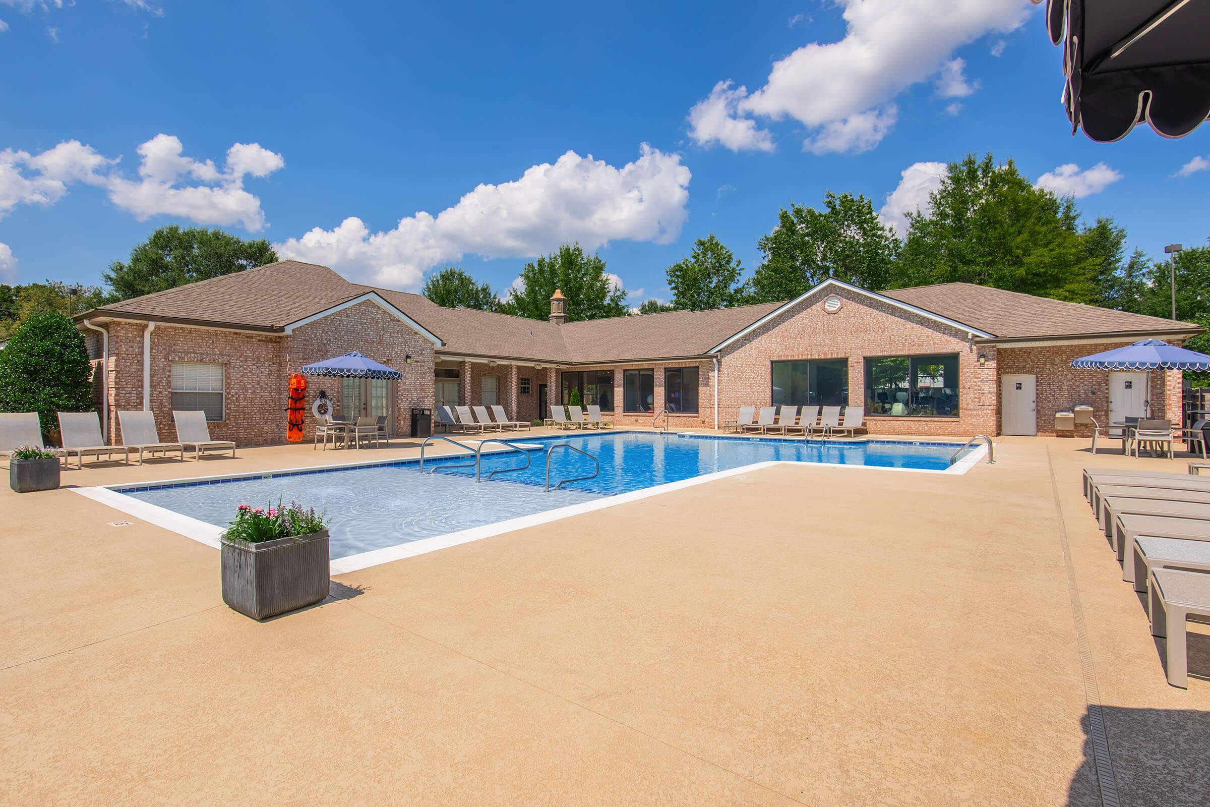 A clear blue sky with fluffy white clouds above a large swimming pool area. The pool is surrounded by lounge chairs and small tables, with a brick building in the background. Umbrellas provide shade, and green trees line the edges, creating a relaxing outdoor atmosphere.