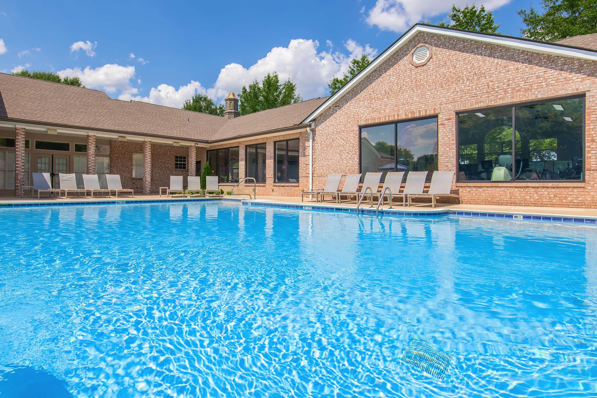 A clear blue swimming pool with sun loungers arranged around it, located next to a brick building with large windows. The sky is bright with fluffy white clouds and lush green trees are visible in the background, creating a relaxing outdoor atmosphere.