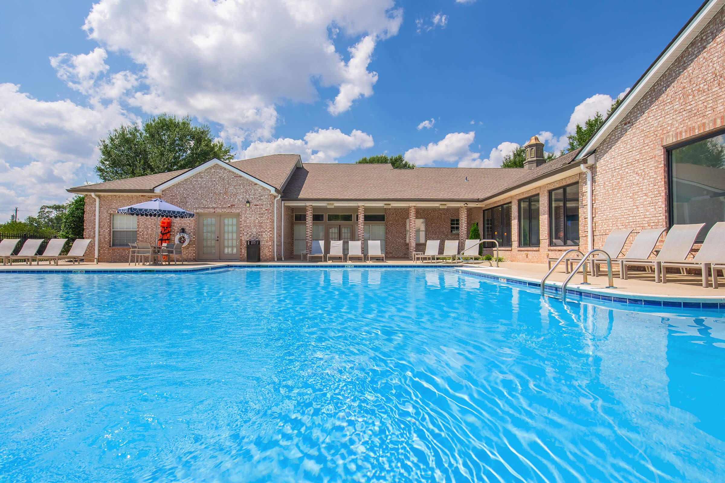 A clear blue swimming pool surrounded by lounge chairs, with a brick building in the background. There are fluffy clouds in a bright sky, and an umbrella near the pool. The setting appears inviting and recreational, ideal for relaxation and leisure activities.