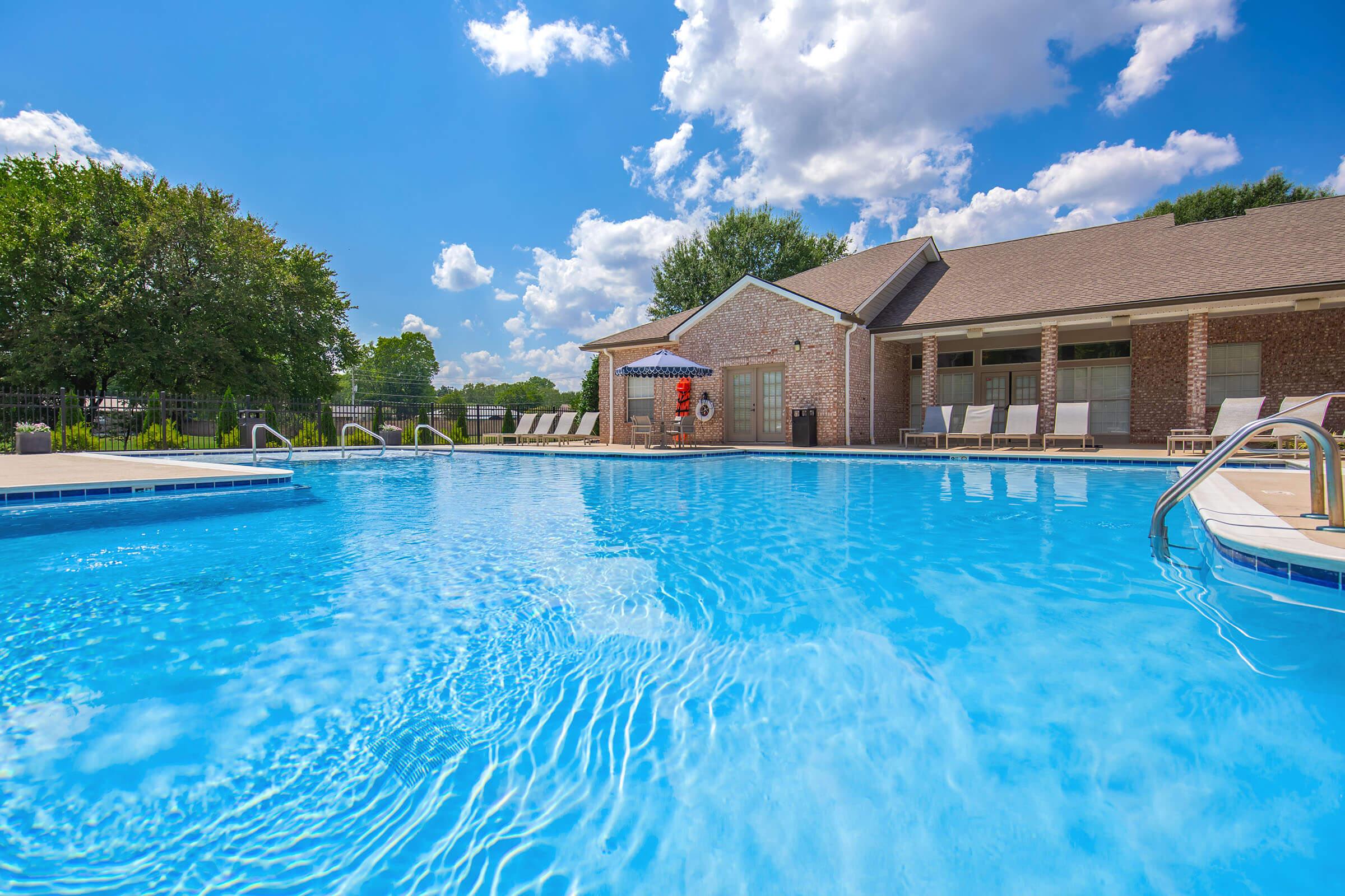 A clear blue swimming pool surrounded by green trees and a well-maintained area. In the background, there is a building with a patio and lounge chairs, under a bright blue sky filled with clouds. The pool features a ladder and a red life preserver nearby, creating a relaxing atmosphere.