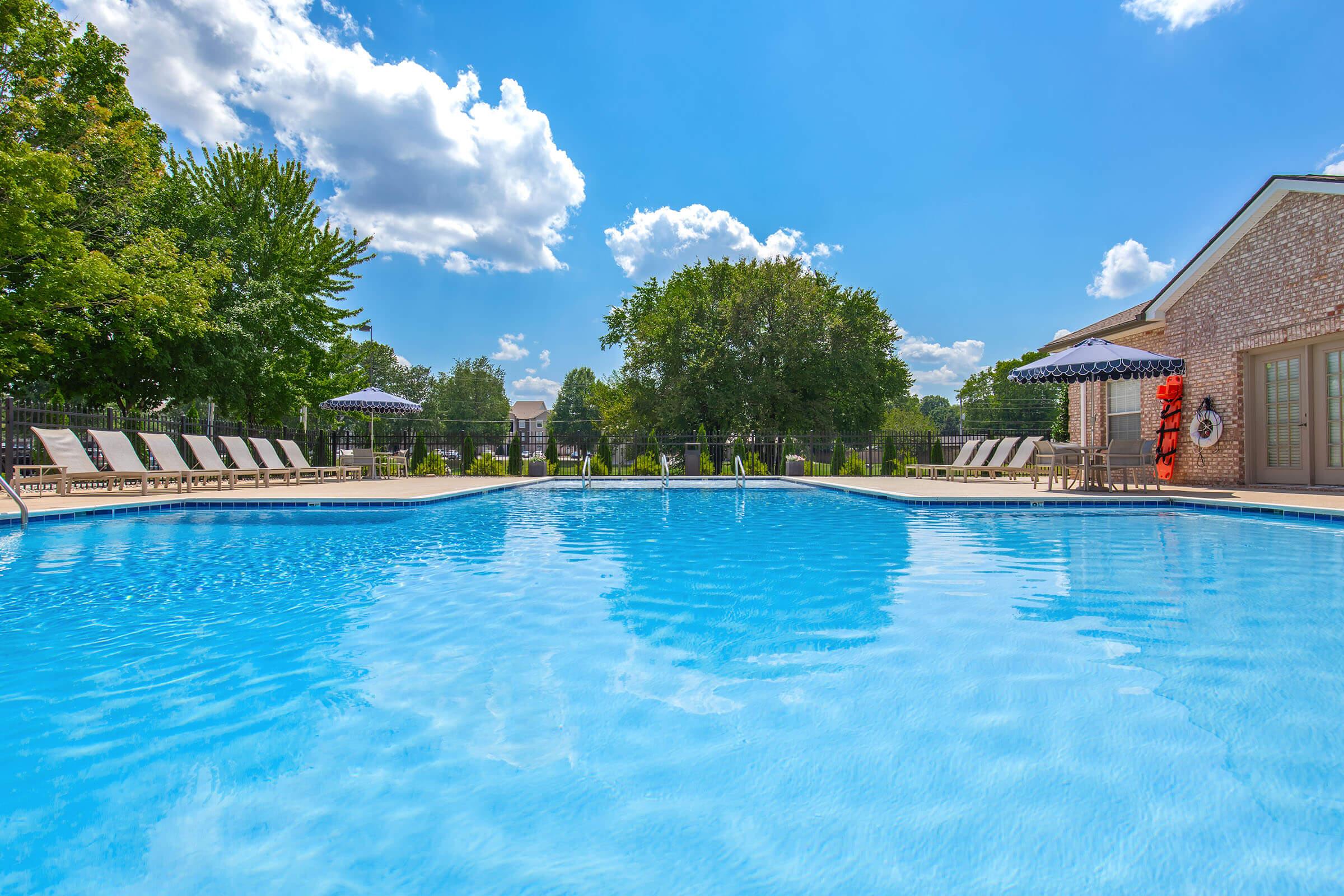 A clear blue swimming pool surrounded by lounge chairs and umbrellas, set against a backdrop of green trees and a bright blue sky with fluffy white clouds. The scene conveys a relaxing outdoor atmosphere, ideal for leisure and summer activities.