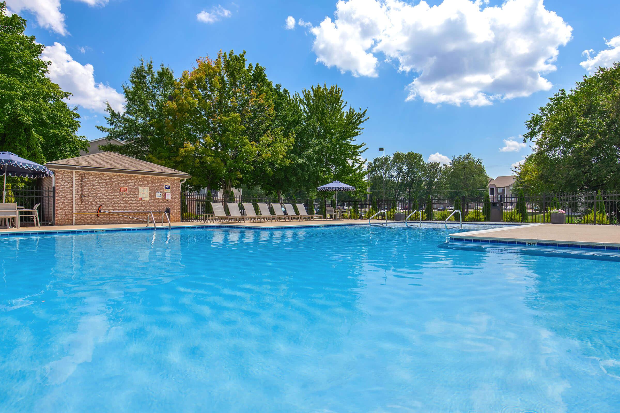 A clear blue swimming pool surrounded by lush green trees and a few shaded lounge chairs. Bright, sunny skies with scattered clouds above. The pool area features a landing with an umbrella, creating a relaxing atmosphere ideal for leisure and enjoyment.