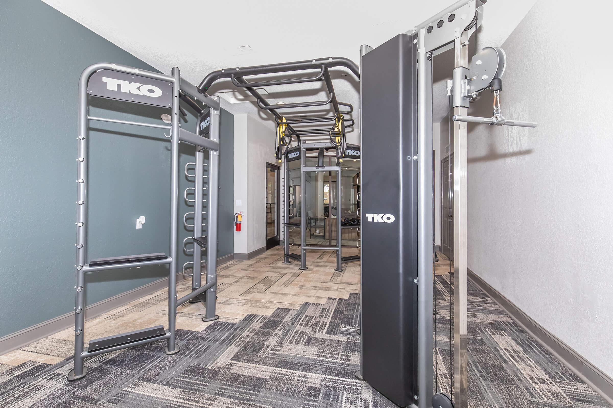 A modern gym space featuring TKO fitness equipment, including a multi-station strength training unit and a pull-up/dip station. The walls are painted in a neutral color, and the floor consists of stylish tiles and carpet. The area is well-lit, creating an inviting atmosphere for workouts.