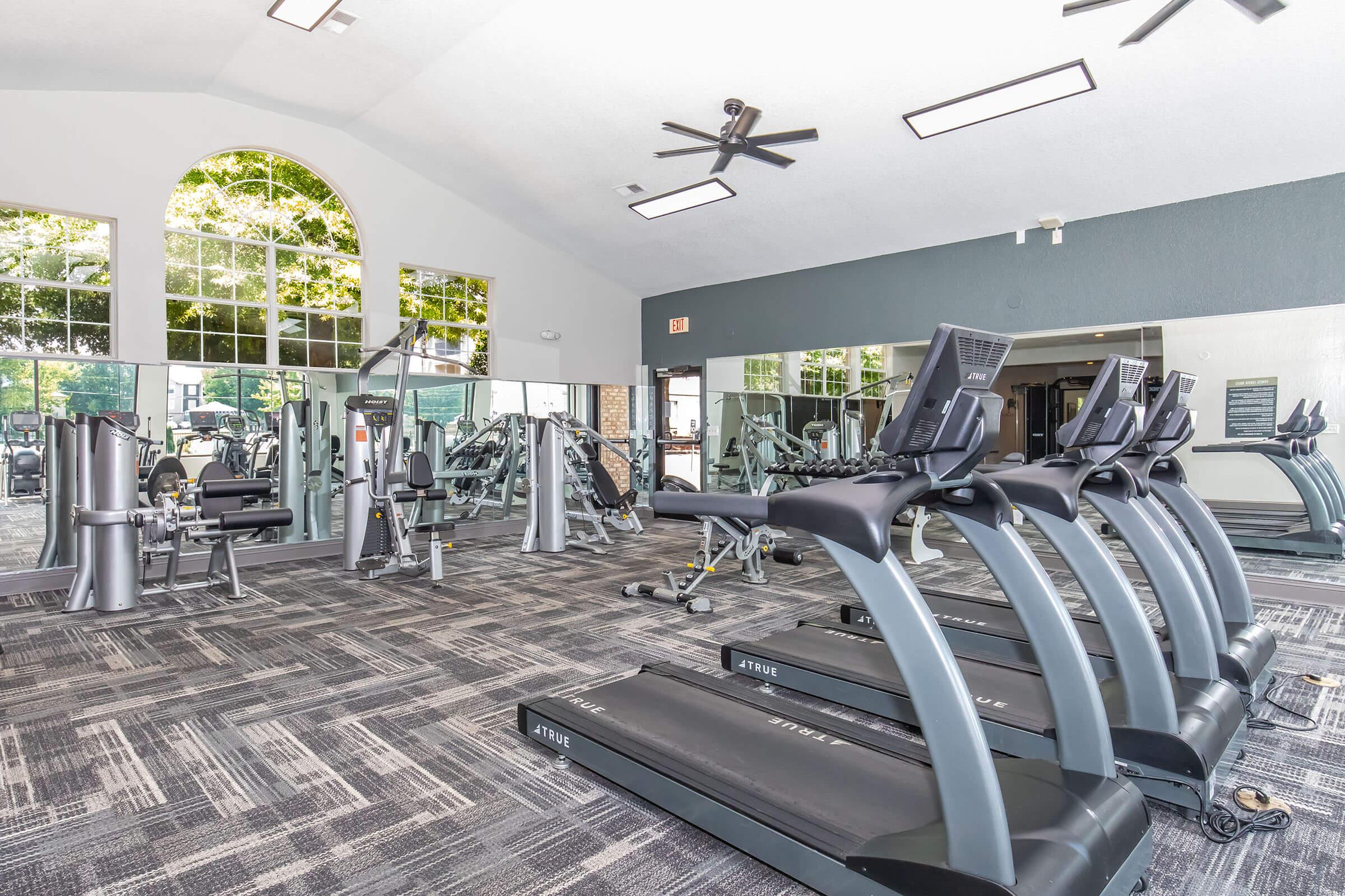 Interior view of a modern gym featuring several treadmills, weight machines, and large windows allowing natural light. The floor is covered with a textured carpet, and there are mirrors on the walls. The space is well-lit with ceiling fans and a high ceiling, creating a spacious and inviting atmosphere for workouts.