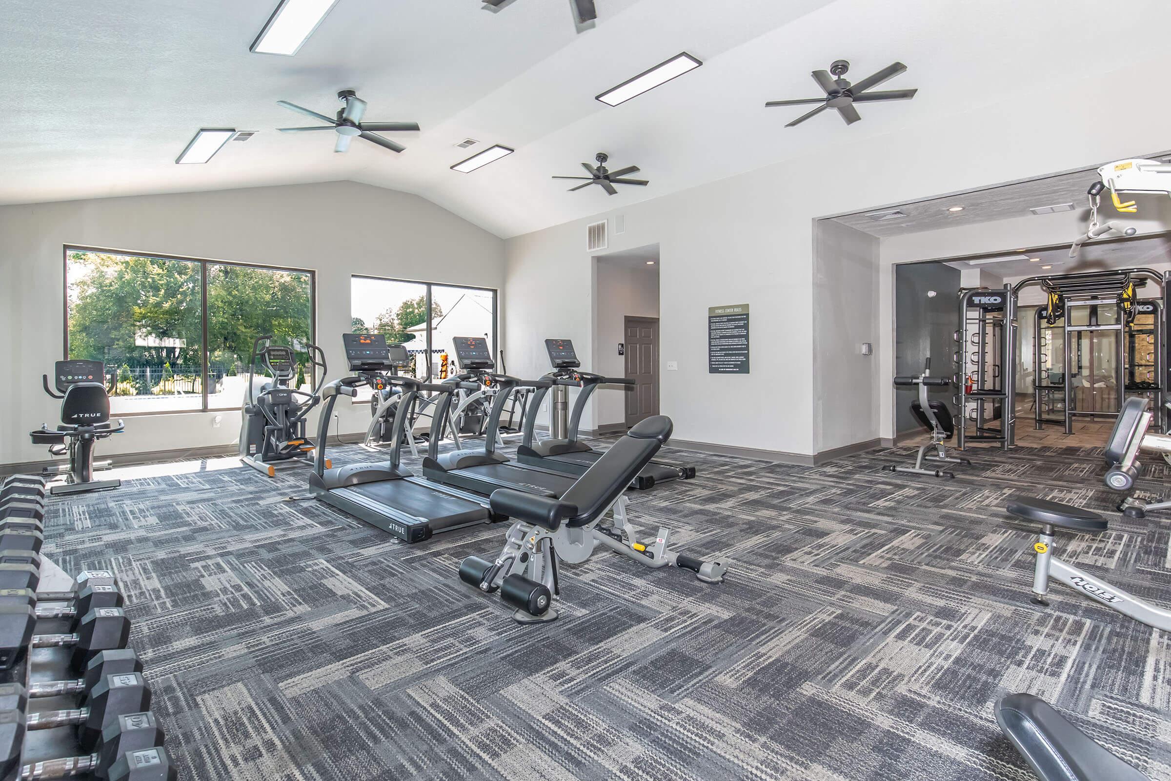 A modern gym interior featuring cardio equipment, weight benches, and a variety of weightlifting machines. Large windows allow natural light in, and ceiling fans are visible. The flooring is a patterned carpet, creating a spacious and inviting workout environment.