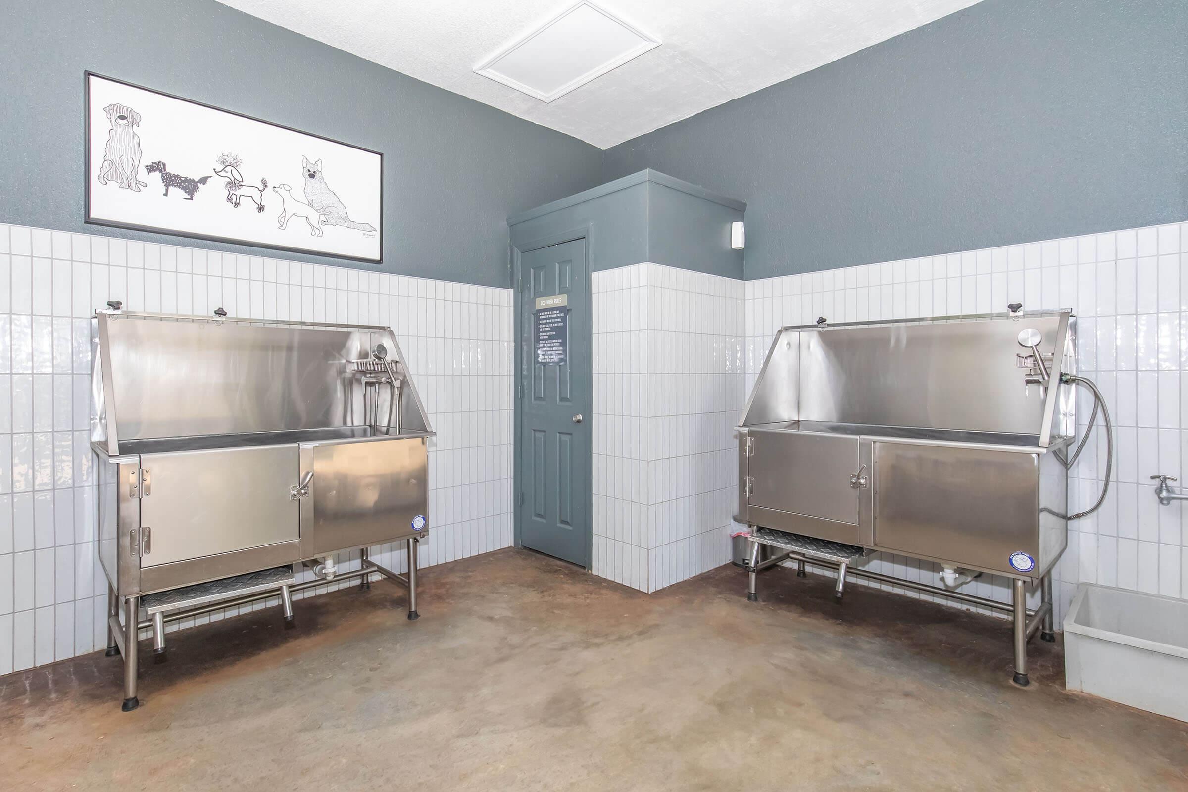 A clean grooming room featuring two stainless steel tubs for pet bathing. The walls are tiled, and there's a framed artwork of playful dogs. A door leads to another area, and the floor is a smooth, light-colored concrete. The space is well-lit and designed for pet care.