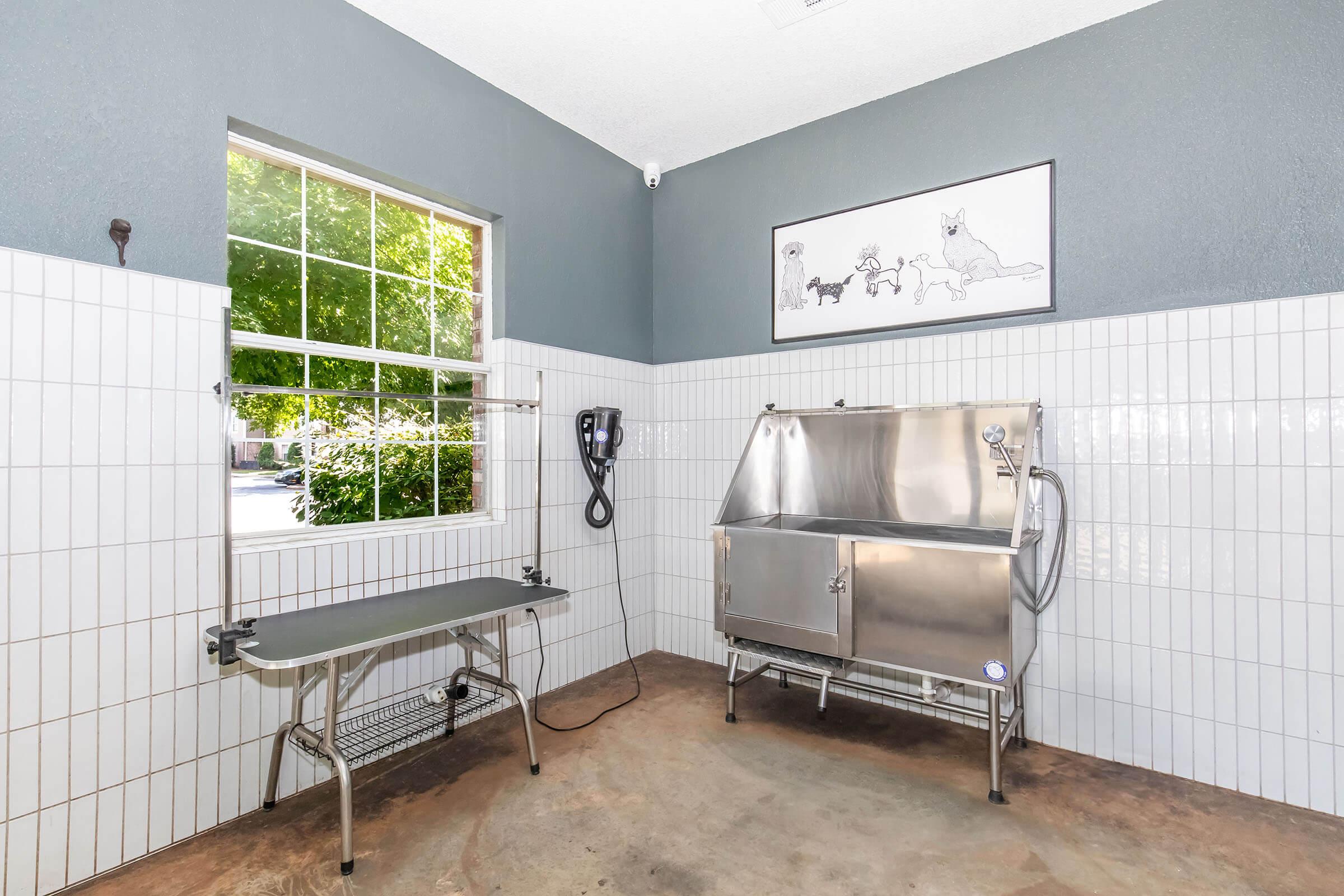 A clean, well-lit grooming room featuring a stainless steel tub for bathing pets, a grooming table with a metal frame, and a wall-mounted pet hair dryer. There is a large window allowing natural light in, adorned with a framed illustration of dogs on the wall. The floor is polished concrete and the walls are tiled halfway up.