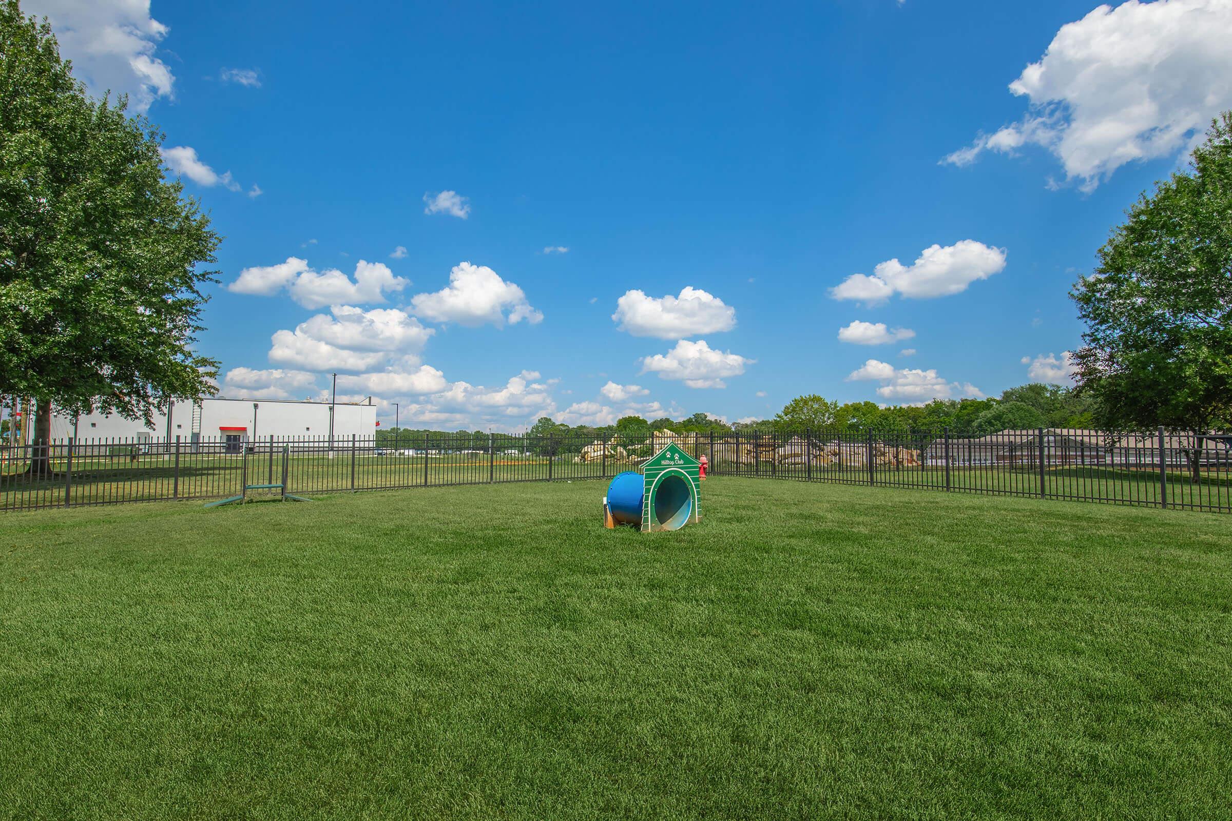 A spacious, well-maintained grassy area enclosed by a fence, featuring a colorful children's play tunnel. The background includes a blue sky with fluffy clouds and trees, along with a building visible in the distance. It's a sunny day, ideal for outdoor play.