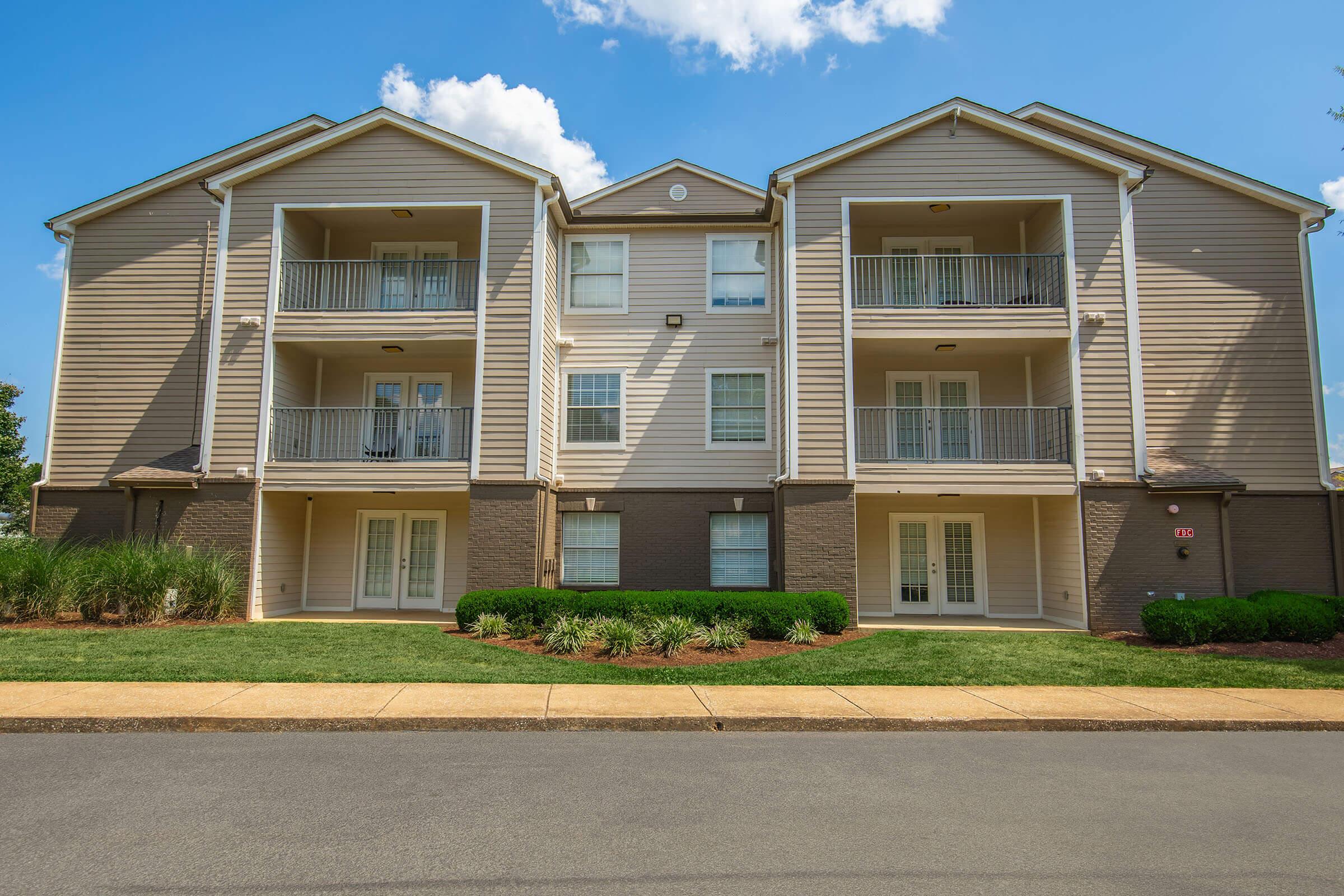 A two-story residential building featuring multiple balconies, light brown siding, and white trim. The front yard is well-maintained with green grass and shrubs, and there is a paved pathway leading up to the entrance. The sky above is blue with scattered clouds.