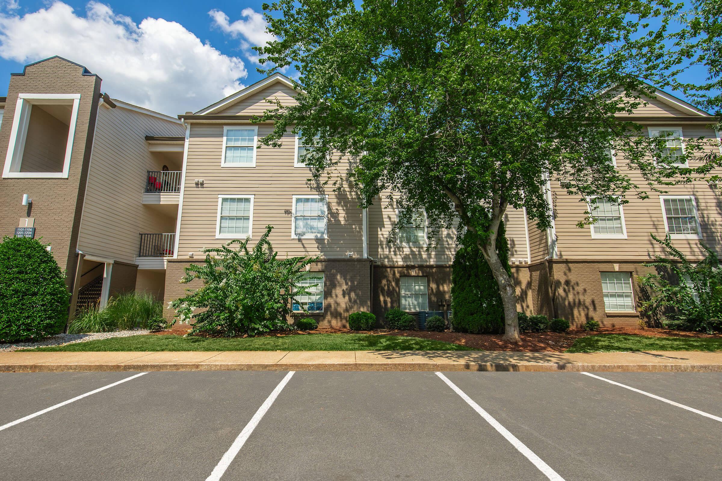 A well-maintained, two-story apartment building with beige siding and large windows, surrounded by greenery and neatly trimmed bushes. A few parking spots are visible in the foreground, and the sky is partly cloudy.