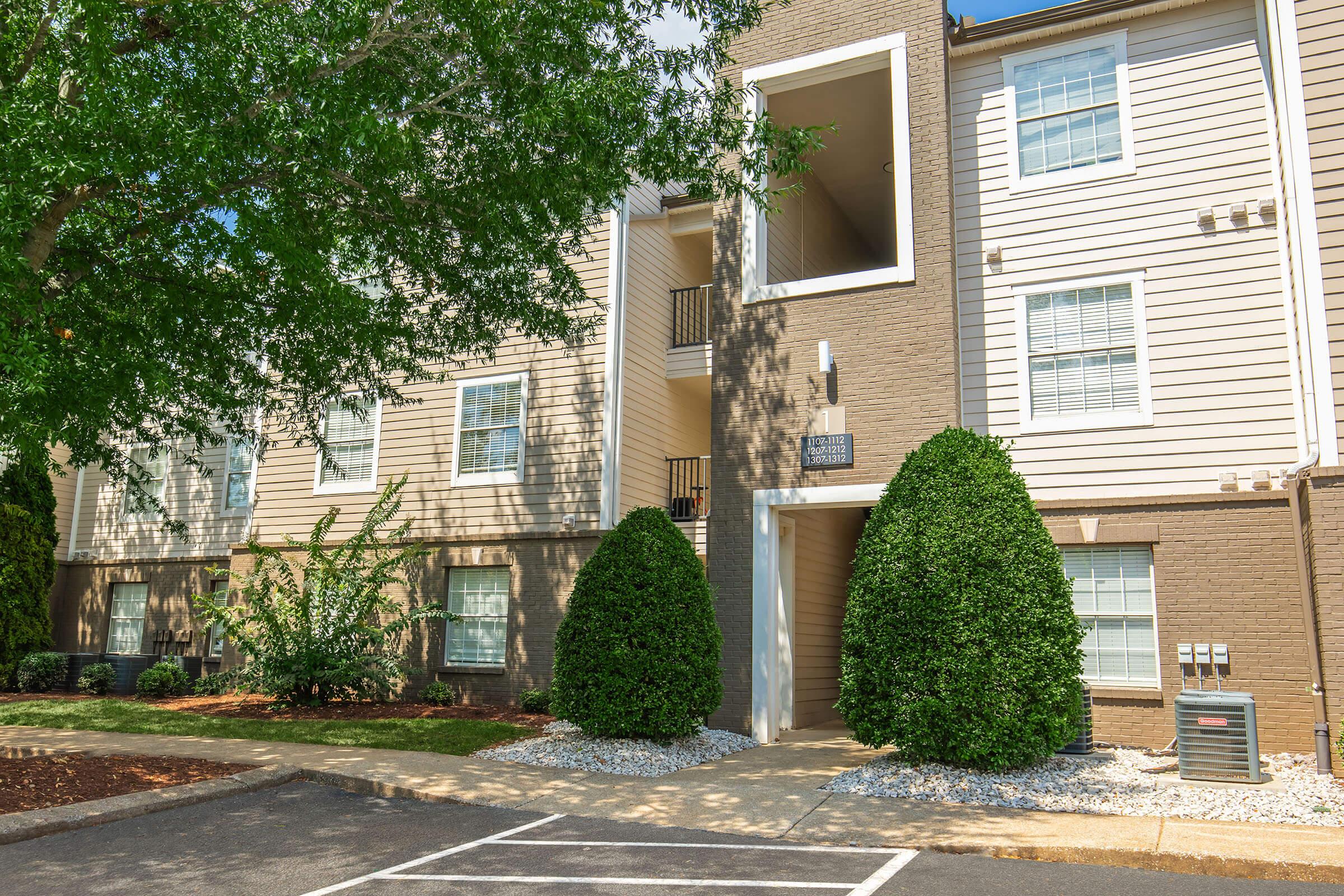 Exterior view of a modern apartment building featuring a tan and brown color scheme. The entrance is framed by two neatly trimmed, green shrubs. The surrounding area has decorative gravel and is bordered by a tree on the left. A parking space in front is visible but empty, suggesting a residential environment.