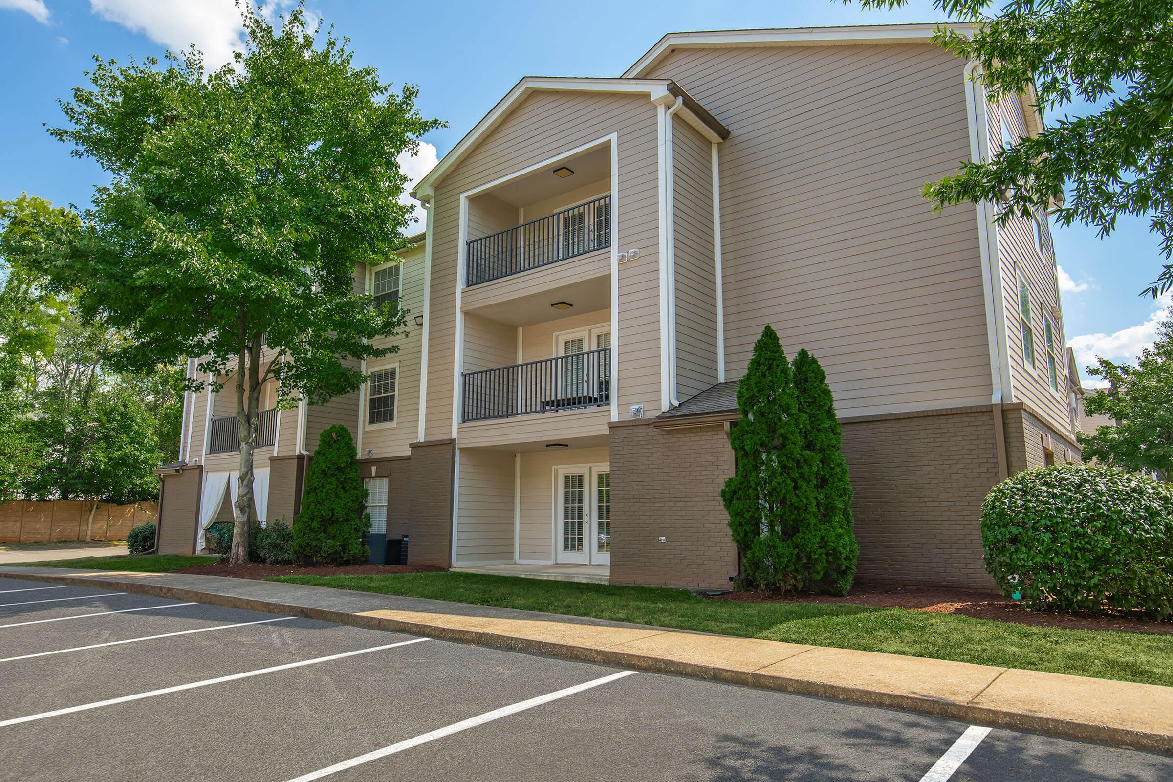 A modern multi-story apartment complex with a light-colored facade, balconies, and well-maintained landscaping. The building is set against a clear blue sky, and a parking area is visible in the foreground, featuring empty parking spaces lined along the driveway.