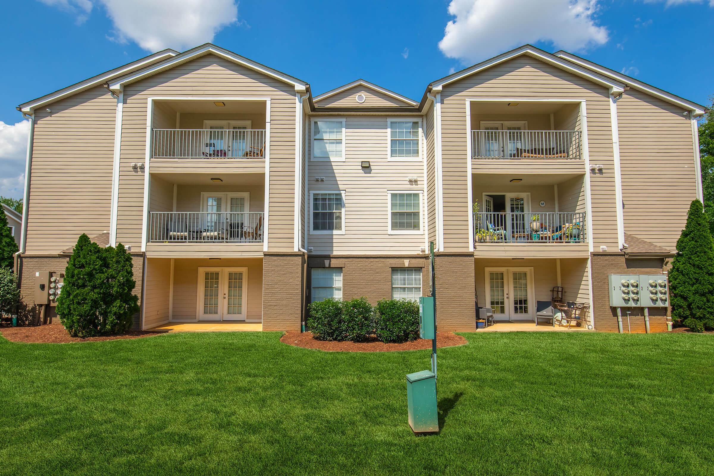 A view of a two-story apartment building with multiple balconies, situated on a well-maintained green lawn. The exterior features beige siding, with decorative plants and seating visible on the balconies. There is a central pathway leading to the entrances, and utility boxes are located on the side.