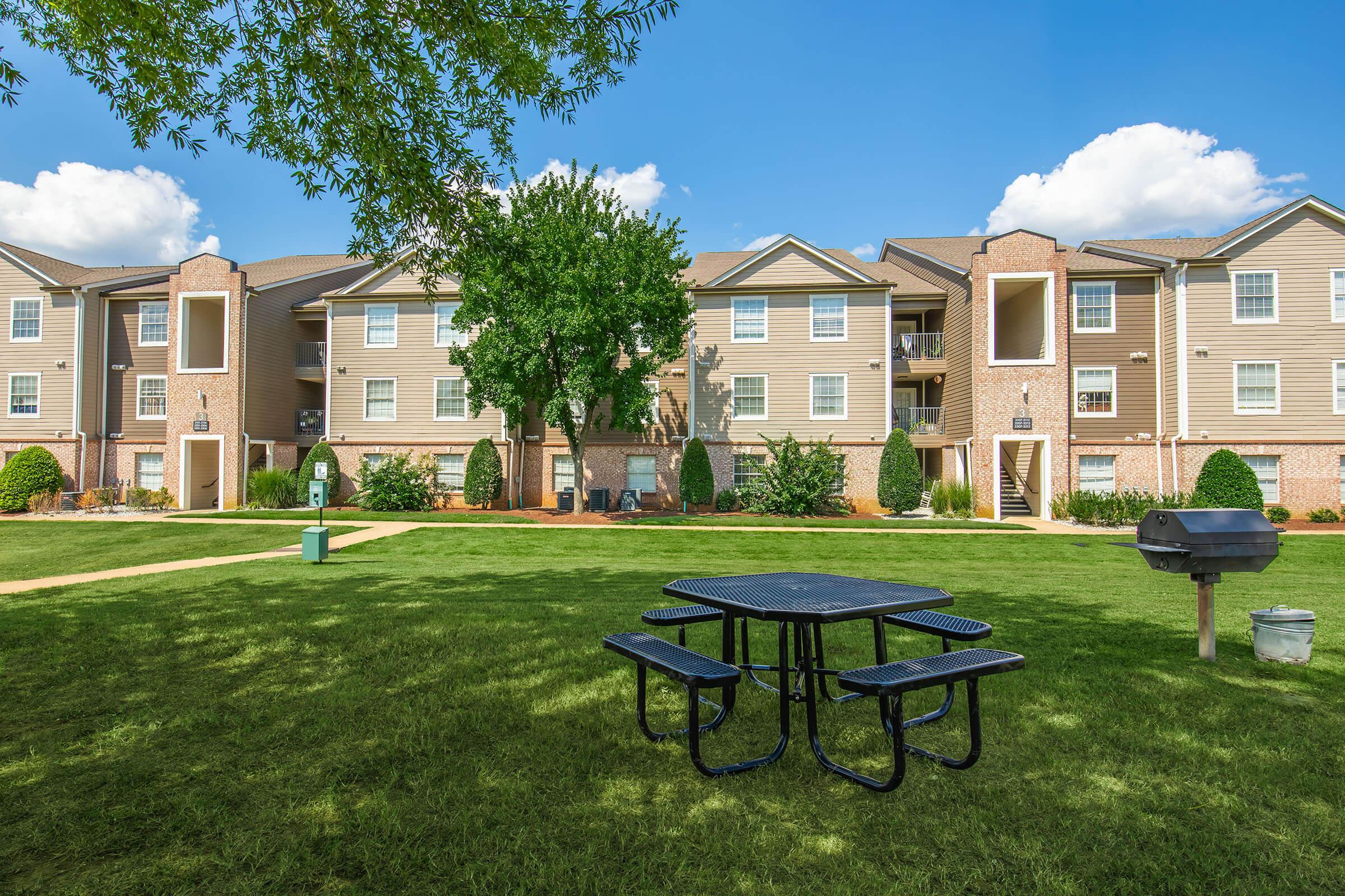 A well-maintained outdoor area with a picnic table and grill, surrounded by green grass and trees. In the background, there are several multi-story apartment buildings with beige siding and landscaped gardens, under a clear blue sky with some clouds.