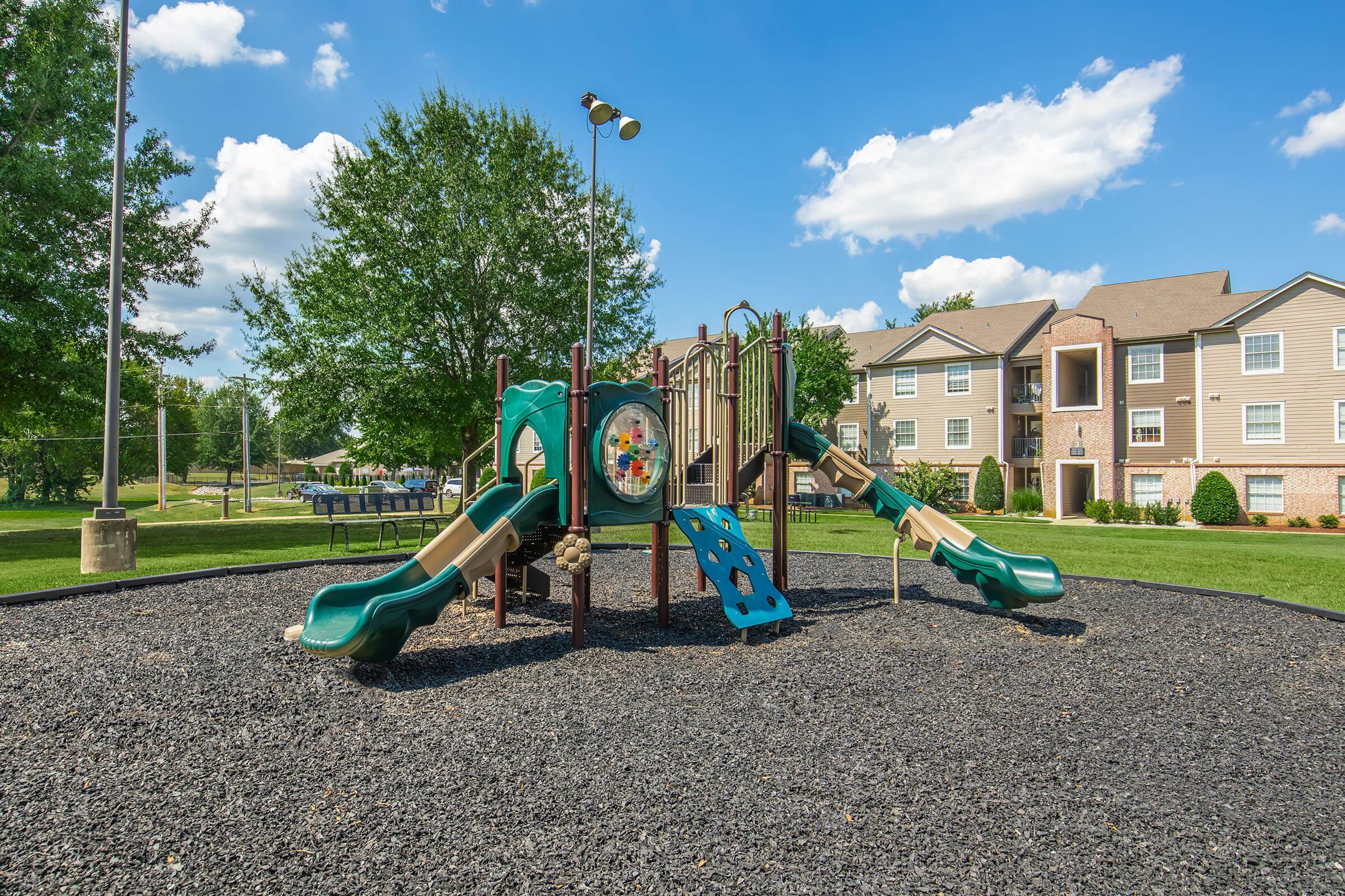 Playground with two green slides and climbing structure, surrounded by black rubber mulch. In the background, there are several residential buildings with green lawns and trees under a blue sky with fluffy white clouds.