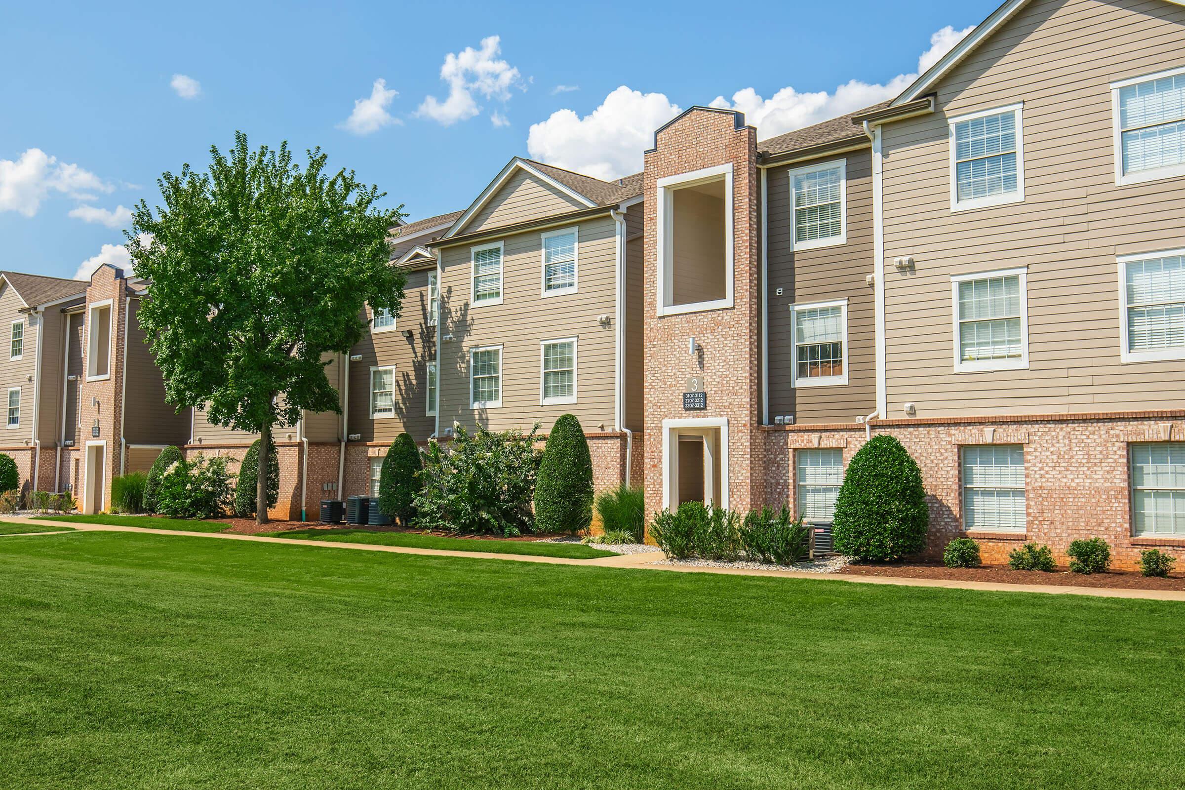 A well-maintained apartment complex featuring multiple two-story buildings with a mix of brick and siding exteriors. Lush green lawns and neatly trimmed shrubs surround the buildings, under a clear blue sky with scattered clouds. The setting appears welcoming and residential.