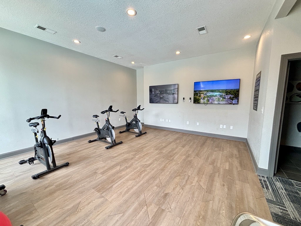 A fitness room featuring several stationary bikes, large windows, and wood-like flooring. Two wall-mounted screens display images, and the space is well-lit with overhead lighting, creating an inviting atmosphere for workouts or cycling classes.