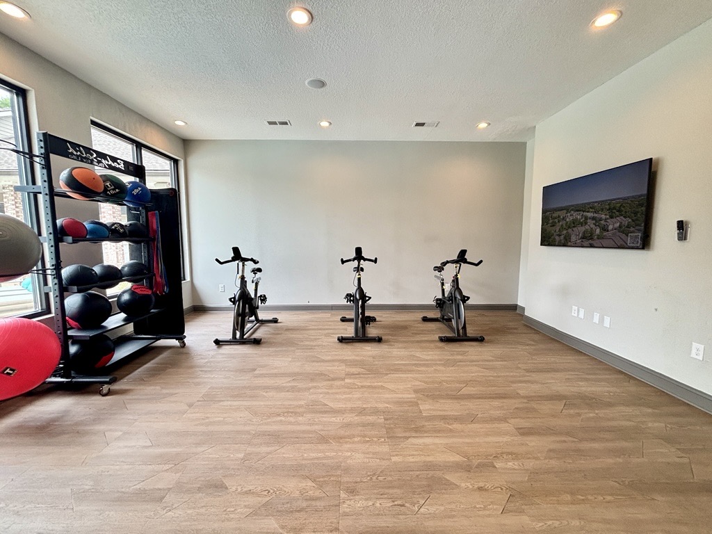 A spacious fitness room featuring three stationary bikes, a wall-mounted TV displaying a scenic view, and a rack with various exercise balls. The room has wooden flooring and recessed lighting, creating a bright and inviting atmosphere for workout activities.