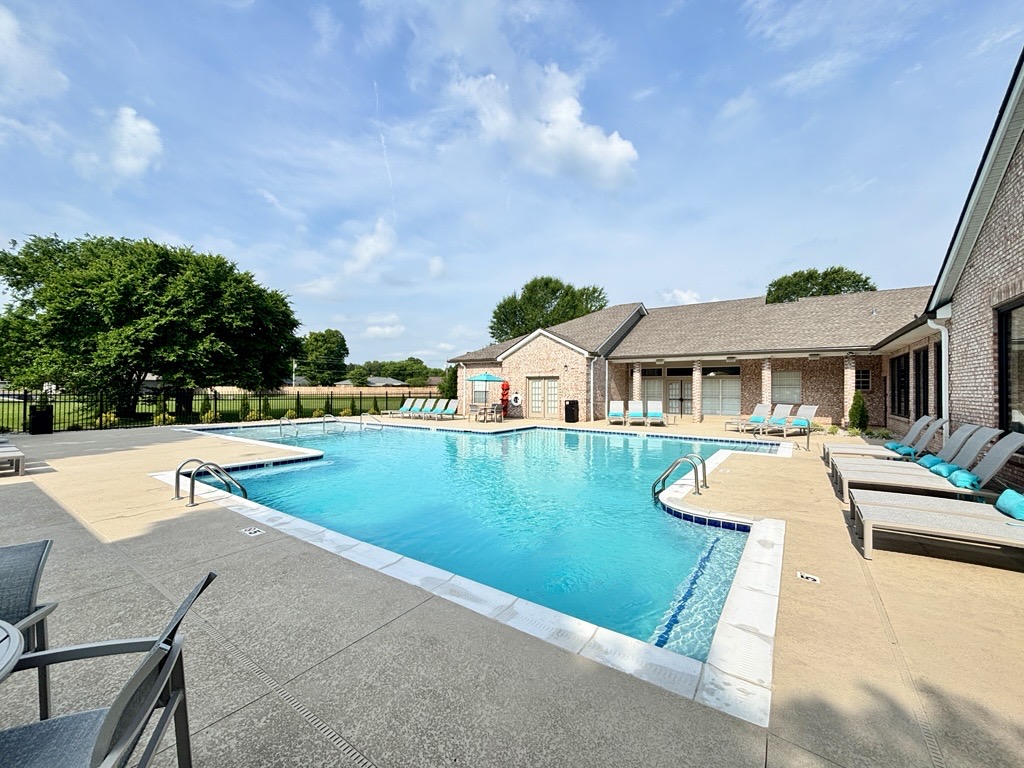 A serene outdoor swimming pool surrounded by lounge chairs, with a fence in the background. The pool area is bathed in sunlight, featuring a clear blue sky and lush green trees nearby. The pool is inviting and well-maintained, offering a relaxing atmosphere for leisure and recreation.
