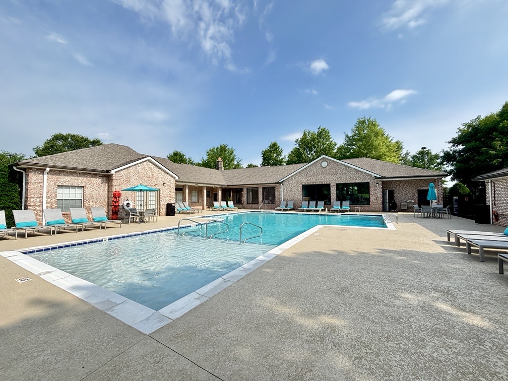 A clear blue swimming pool surrounded by a patio with lounge chairs, umbrellas, and trees. In the background, a brick building with large windows offers access to the pool area. The sky is partly cloudy, creating a serene atmosphere.