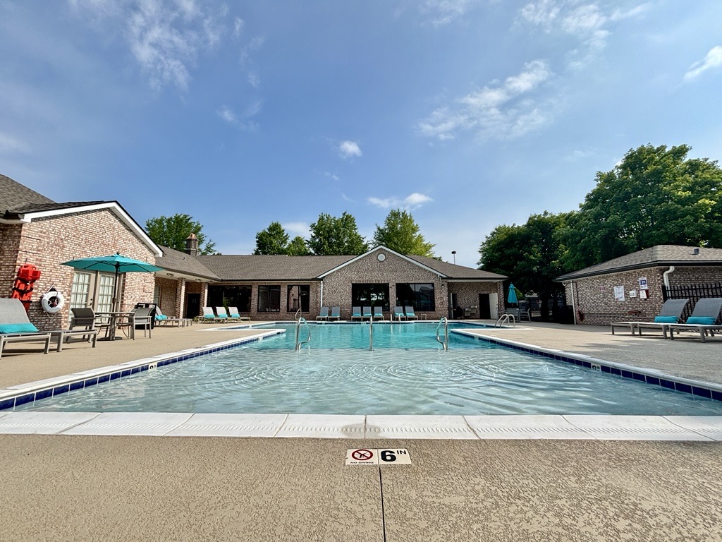A clear swimming pool with a shallow end marked "6 ft" in front of a brick building. The area is surrounded by lounge chairs and umbrellas. Green trees and a blue sky with clouds are visible in the background, creating a relaxing outdoor atmosphere.