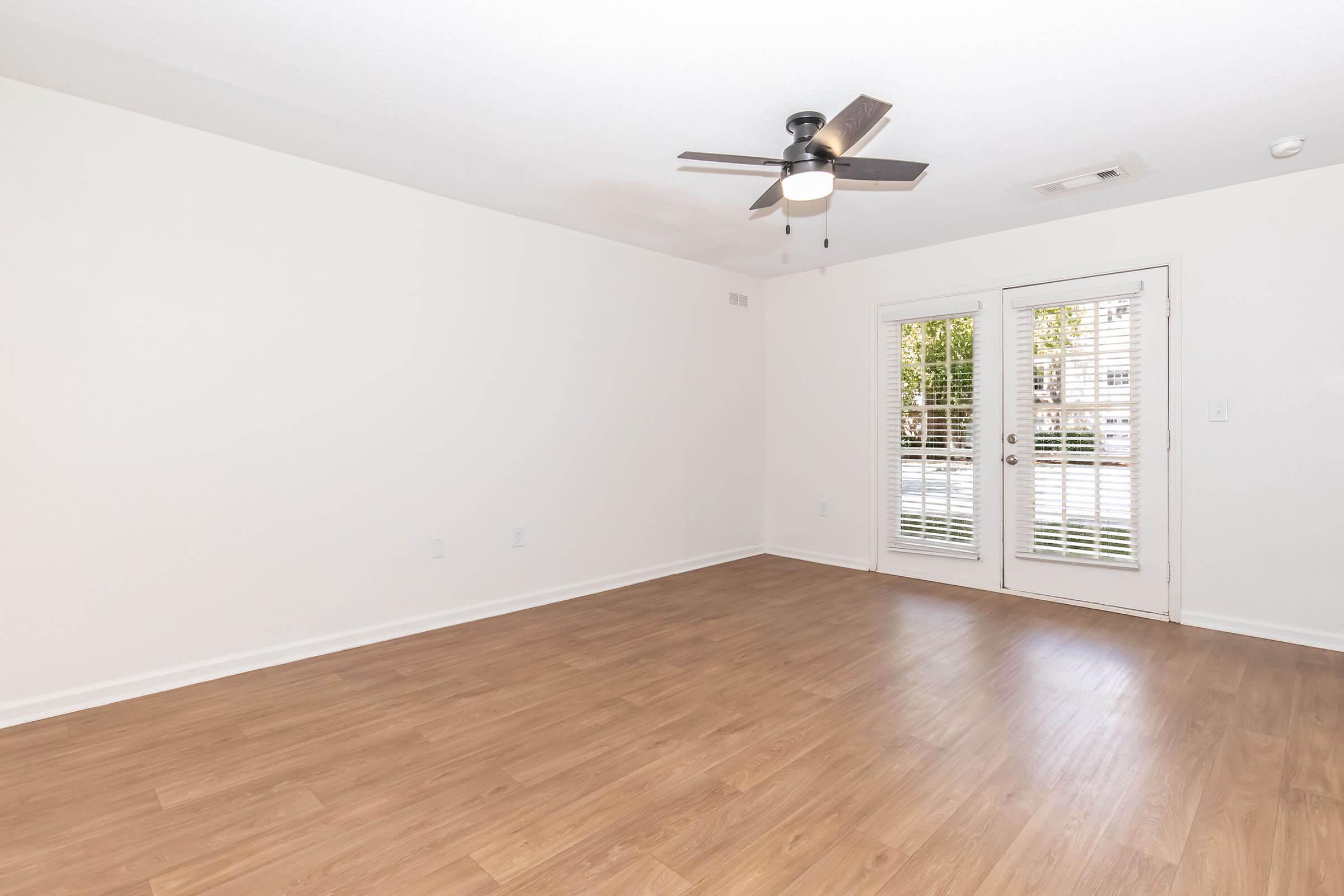 A spacious, empty room with light-colored walls and wooden flooring. It features a ceiling fan and two glass doors that lead to an outdoor area, with natural light coming through. The room is designed for versatility in use and decor.