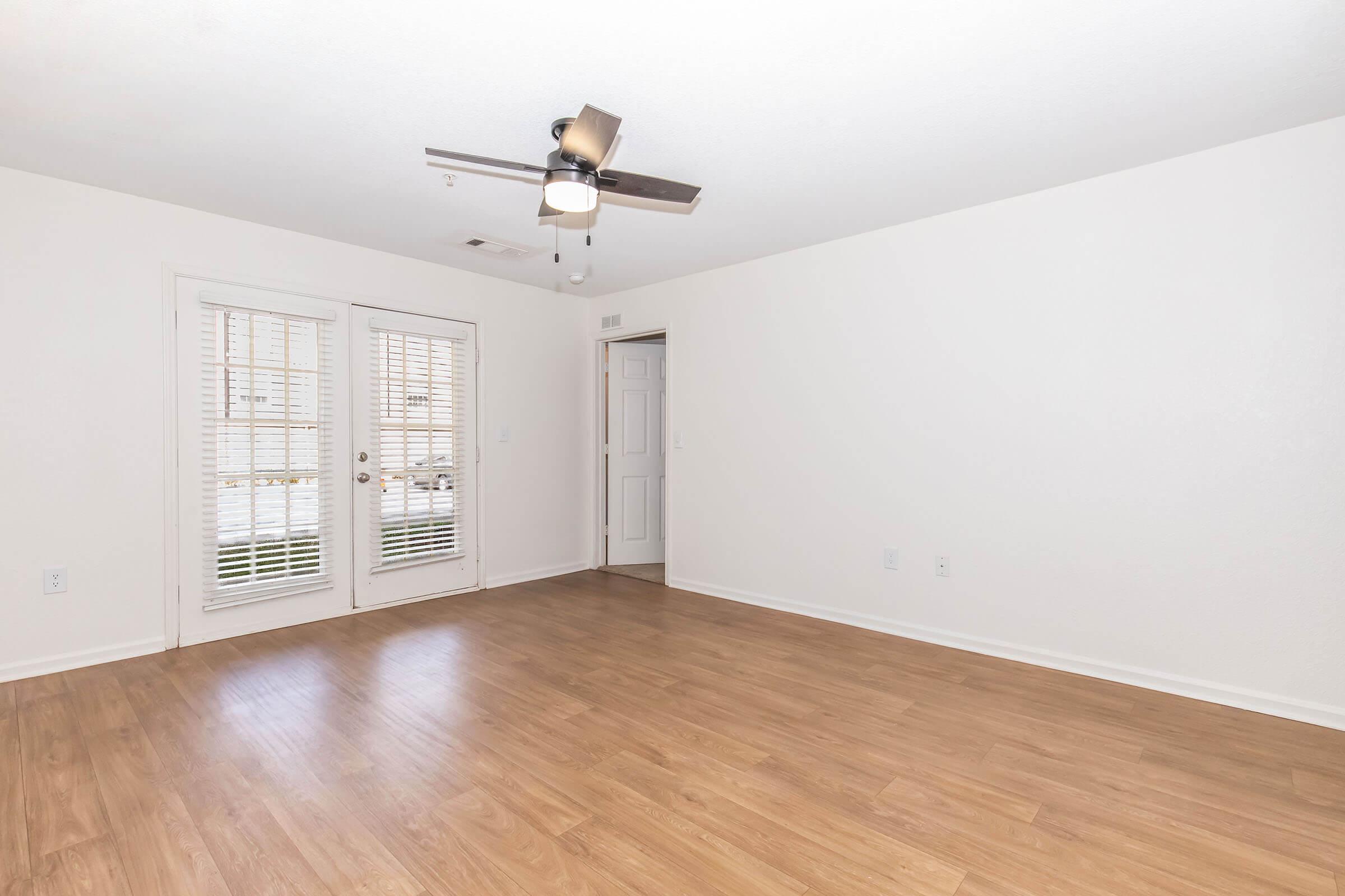 A bright and spacious room with light-colored walls and wooden flooring. There are double glass doors leading outside, and a ceiling fan is installed. A door on the right likely leads to another room. The overall ambiance is modern and inviting, suitable for various uses.