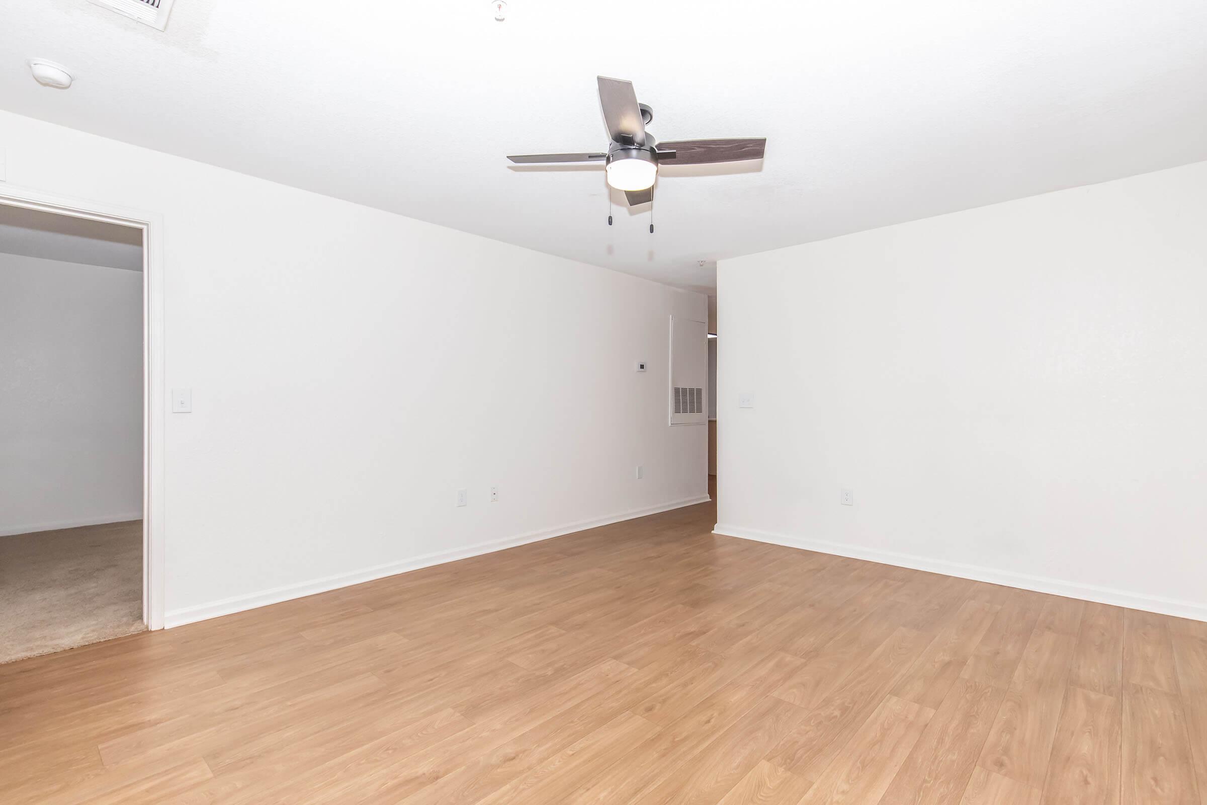 A spacious, empty room with light wooden flooring and white walls. A ceiling fan is visible in the center, and an open doorway leads to another room. The space is well-lit, creating a bright and airy atmosphere.
