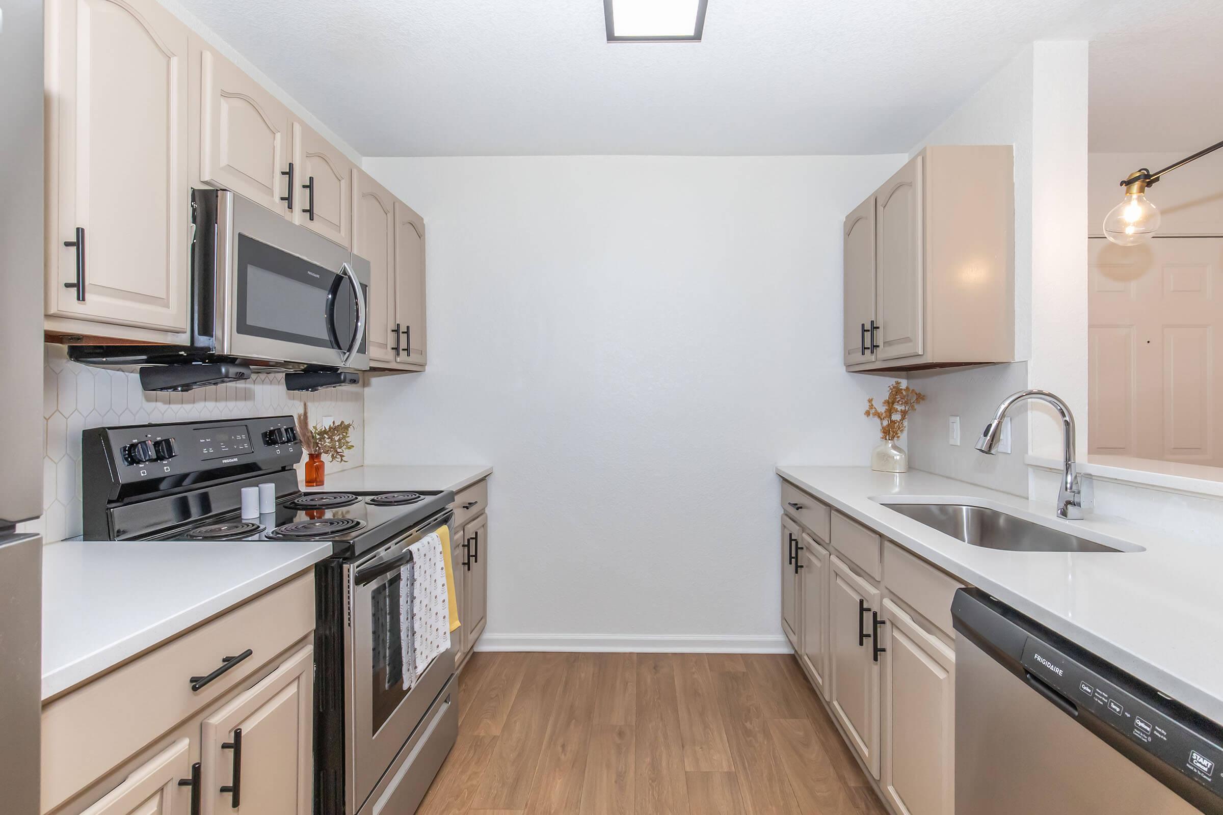 Modern kitchen with light-colored cabinetry, stainless steel appliances including a microwave and oven, and a sleek countertop. The room features a neutral color palette, wooden flooring, and a stylish sink. Decor elements include a decorative vase on the counter, creating a warm and inviting atmosphere.