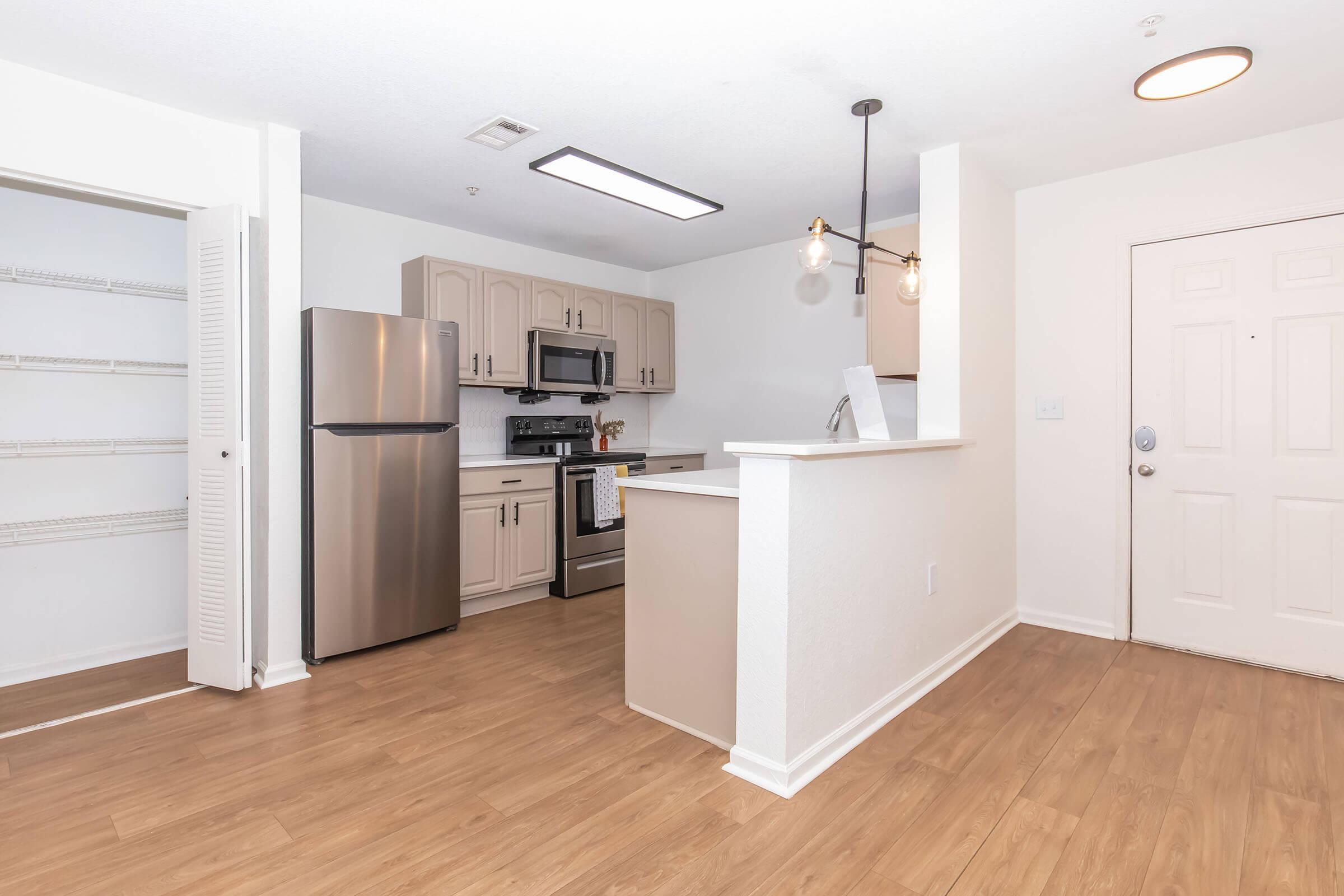 A modern kitchen featuring stainless steel appliances, including a refrigerator and stove. The light wood floor complements the beige cabinets. A simple island separates the kitchen from the entryway, which includes a front door and an open shelving area. The room is well-lit with natural light.