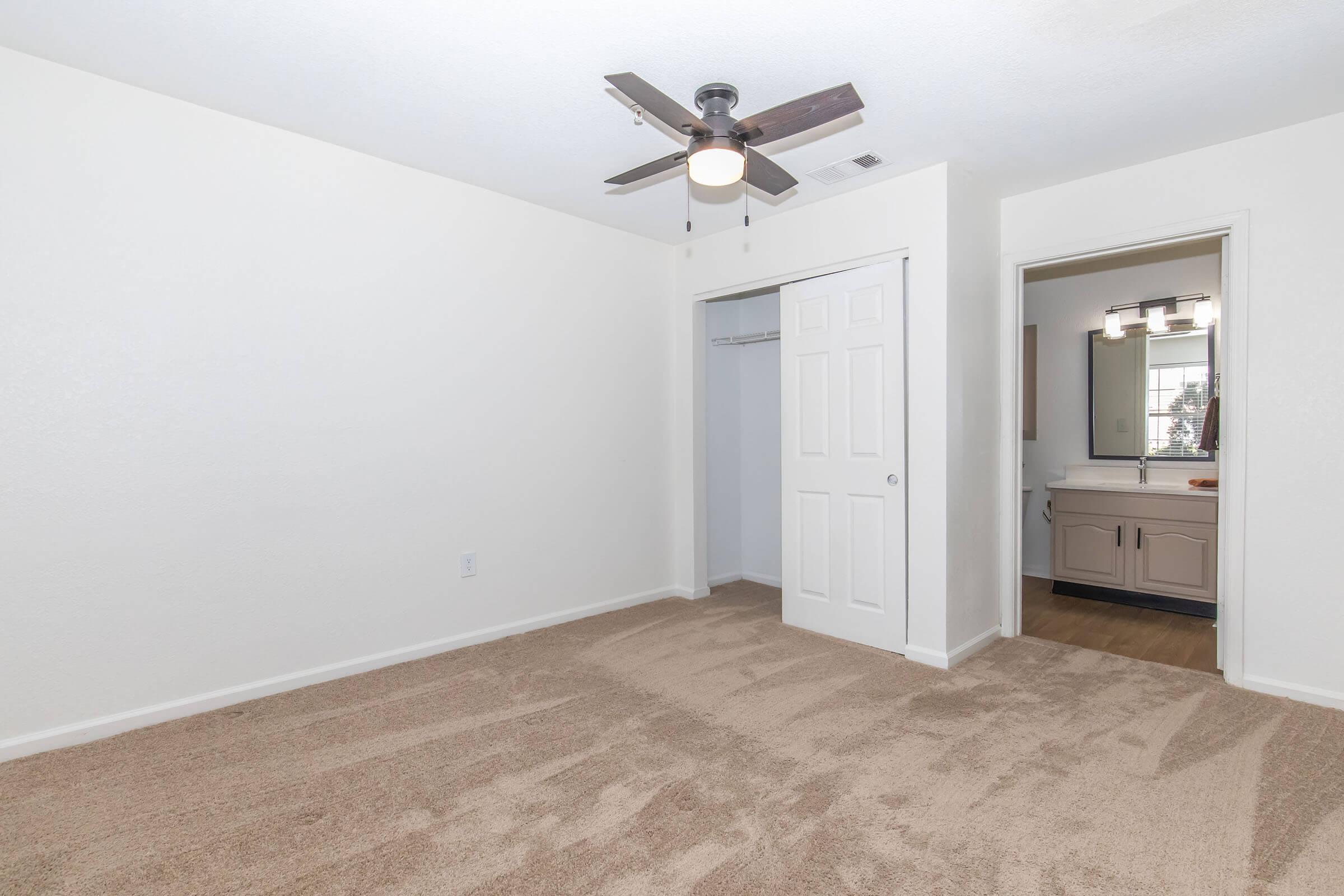 A well-lit, empty bedroom featuring beige carpeting and white walls. The room includes a ceiling fan, a closet with a white door, and a doorway leading to a bathroom area with a mirror and sink visible. The overall appearance is clean and spacious, suitable for decorating.