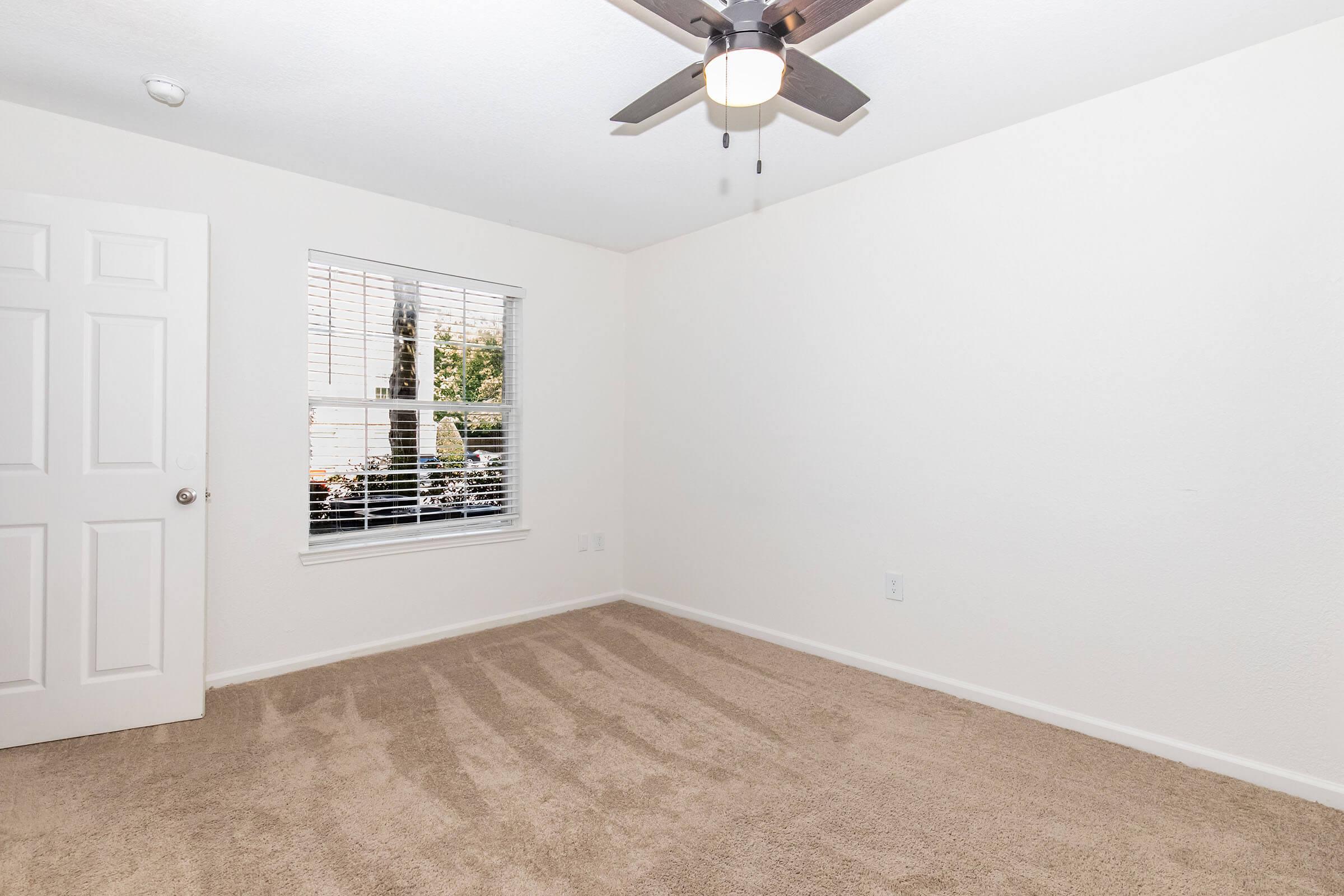 Empty room with light-colored walls and carpet. A window with blinds lets in natural light, and a ceiling fan is mounted in the center. A closed door is visible on the left side of the image, and the overall atmosphere is bright and spacious.