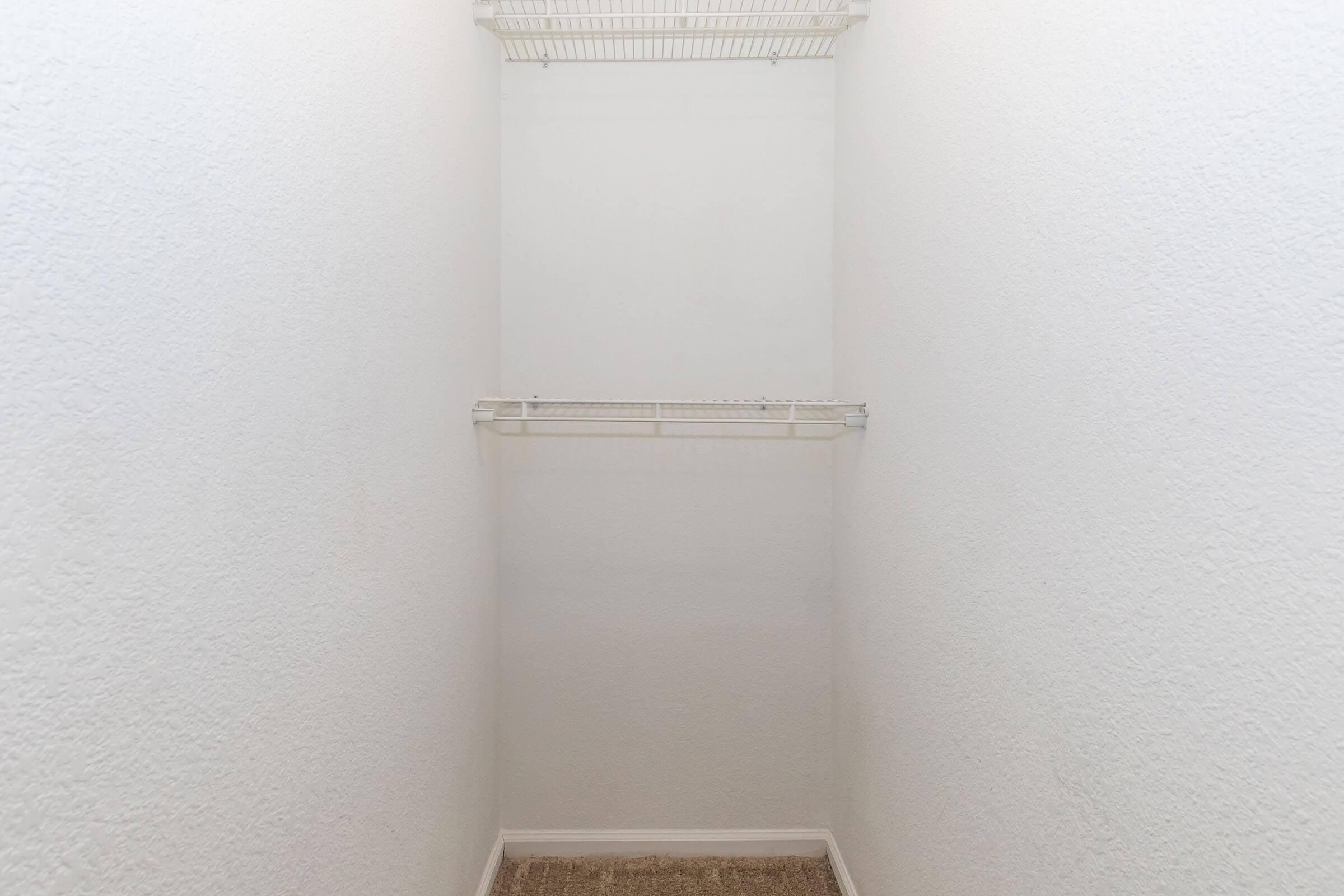 A clean, empty closet with white walls and a wire shelf. The floor is carpeted in a beige color, creating a minimalist and tidy storage space.