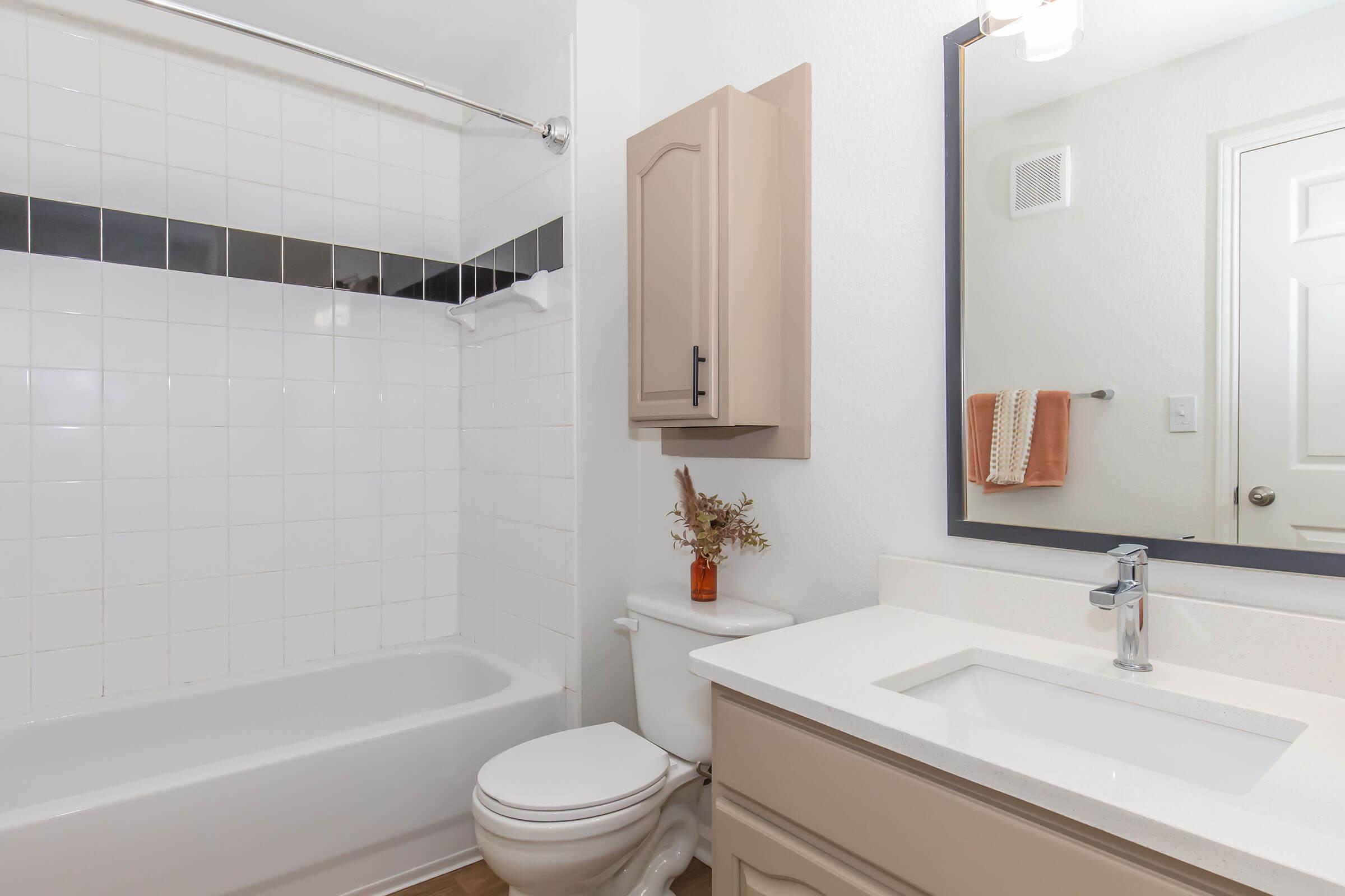A clean and modern bathroom featuring a white tiled bathtub with a shower curtain, a stylish mirror above a sink with a minimalist faucet, and a beige cabinet. There's a decorative flower arrangement on the counter and a towel rack with a towel. The walls are painted light and the space is well-lit.